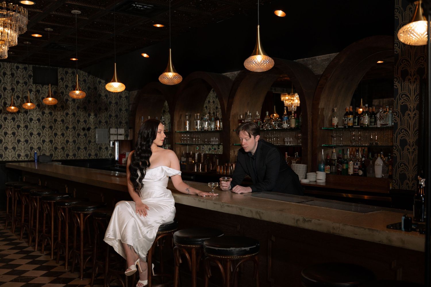 Bride in white dress sits at elegant bar chatting with groom in black suit, romantic dimly lit venue.