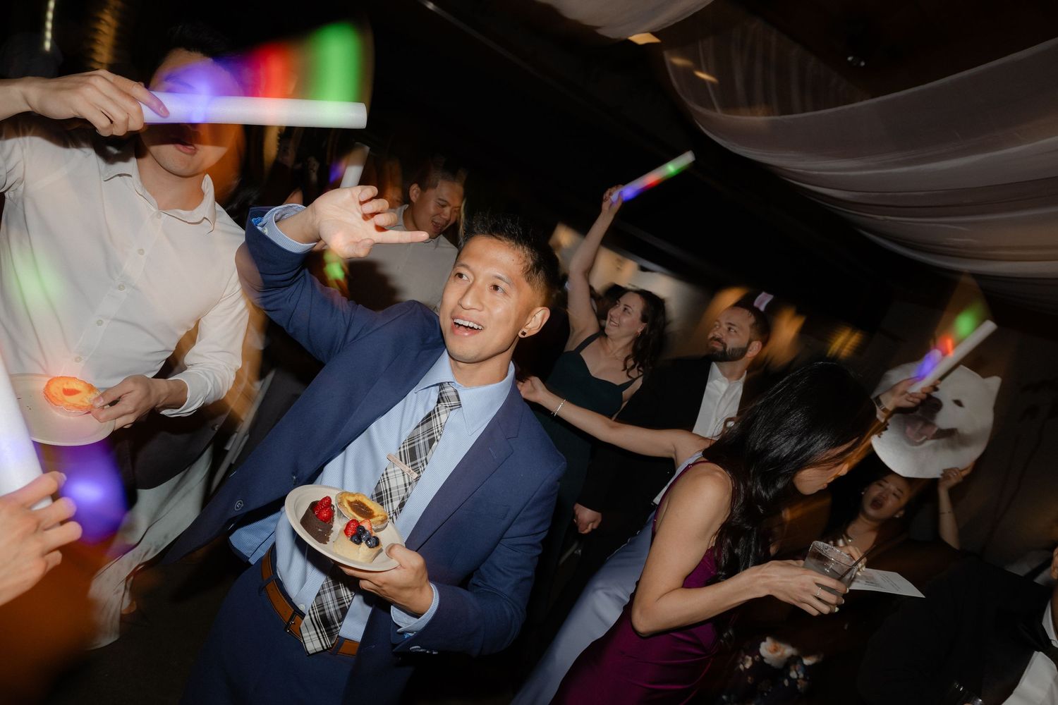 Smiling man in blue suit holding a colorful cake slice while guests dance with glow sticks.