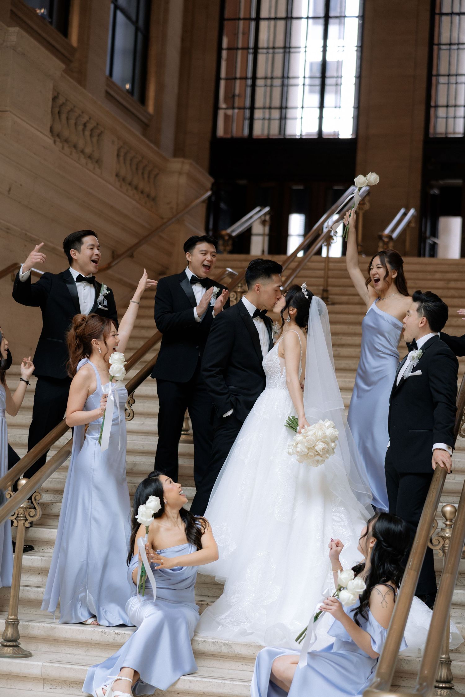 Joyful wedding party celebrates on grand staircase with bridesmaids in blue gowns.