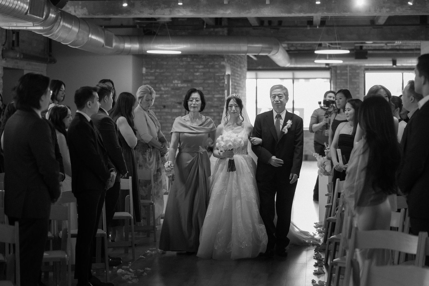 Black and white photo of a bride walking down the aisle with her parents during a wedding ceremony.