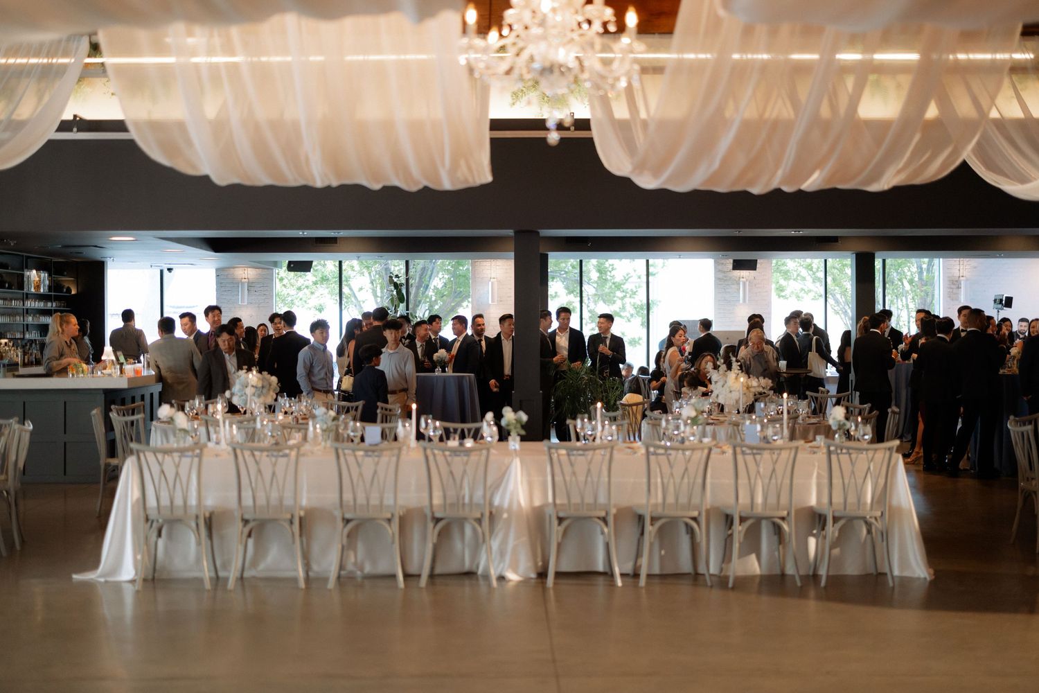 Elegant wedding reception hall with white draped ceilings, chandelier, long banquet tables, and guests mingling in a modern venue.