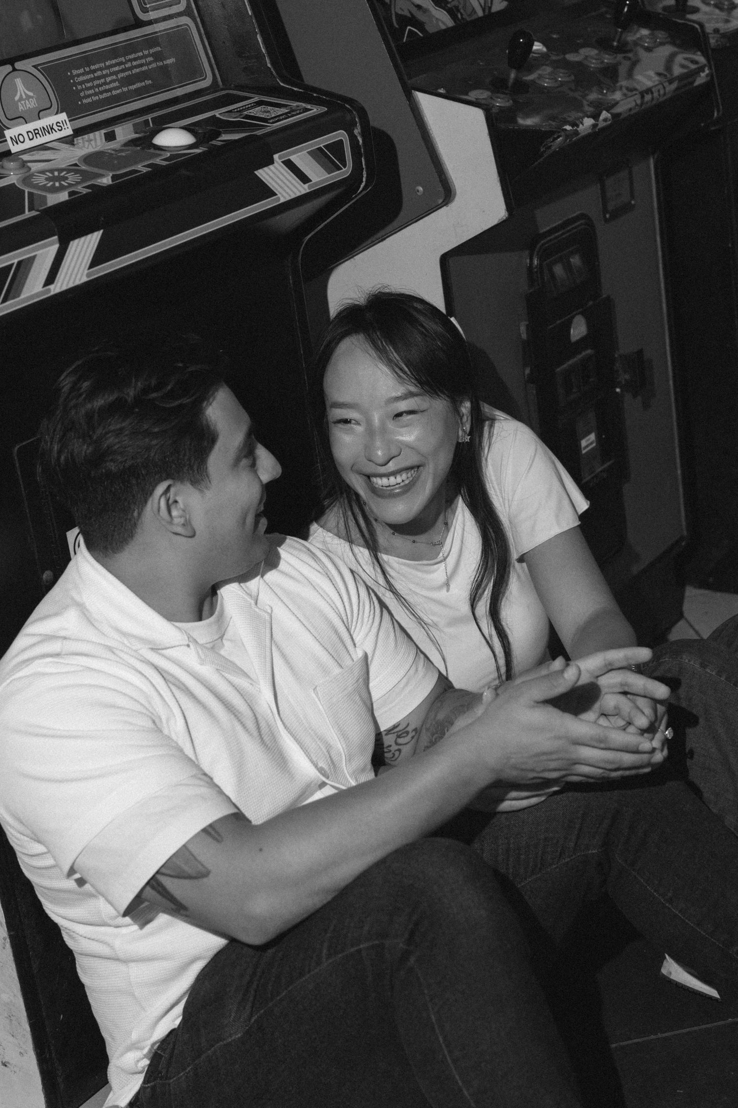 Black and white photo of a couple laughing together while sitting near vintage arcade games.