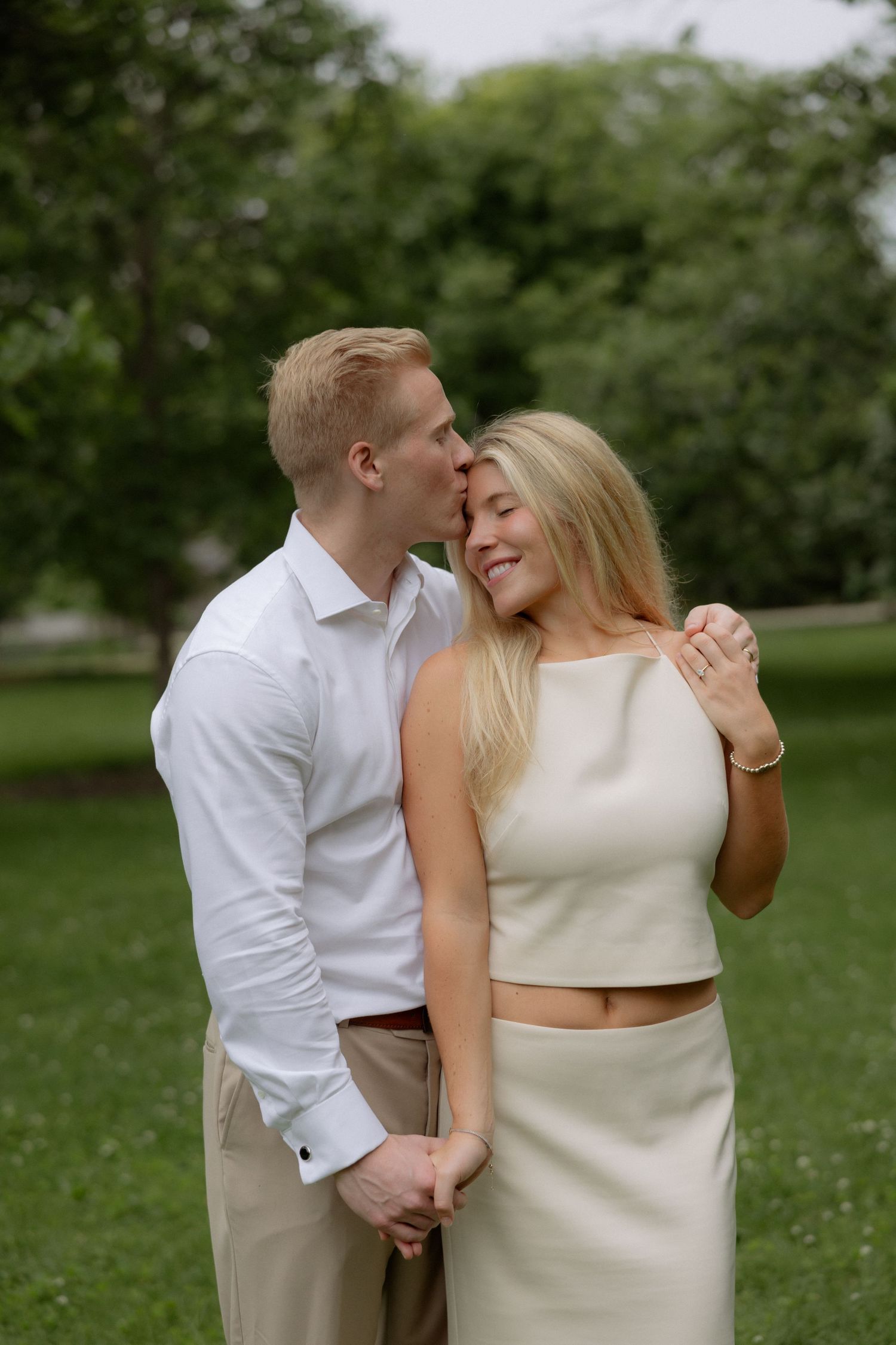 A man in white shirt kisses a smiling blonde woman on the cheek while holding hands in a green park.