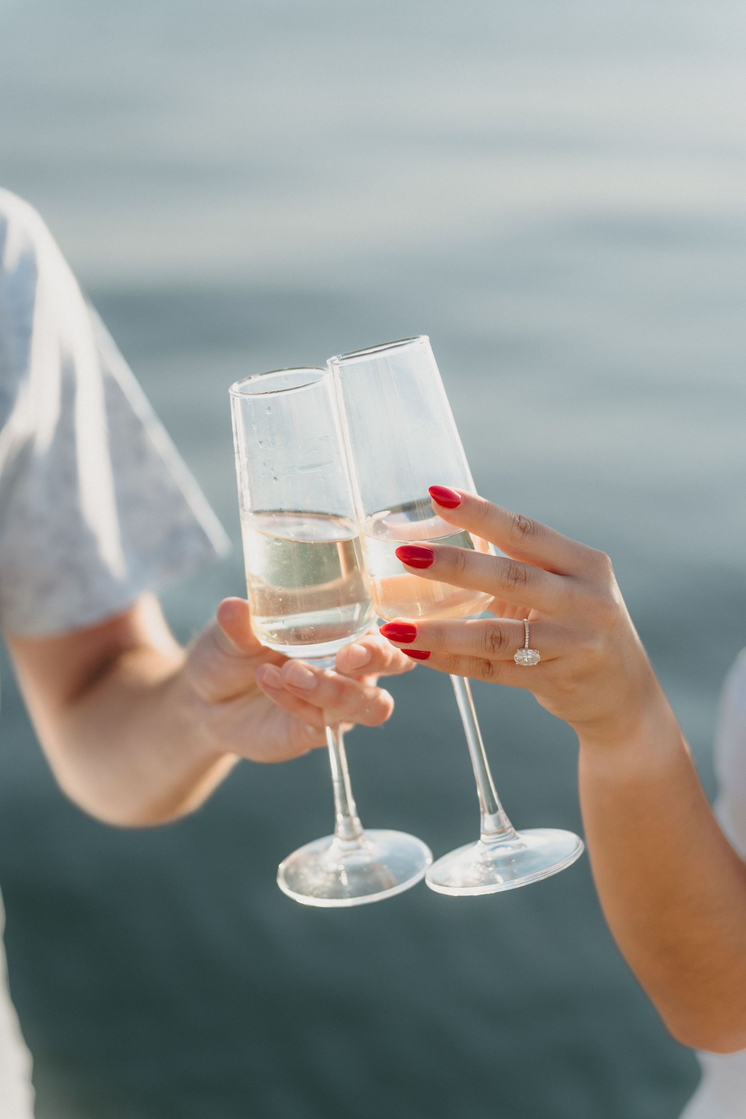 Two hands clinking champagne flutes near water, one wearing a diamond engagement ring.