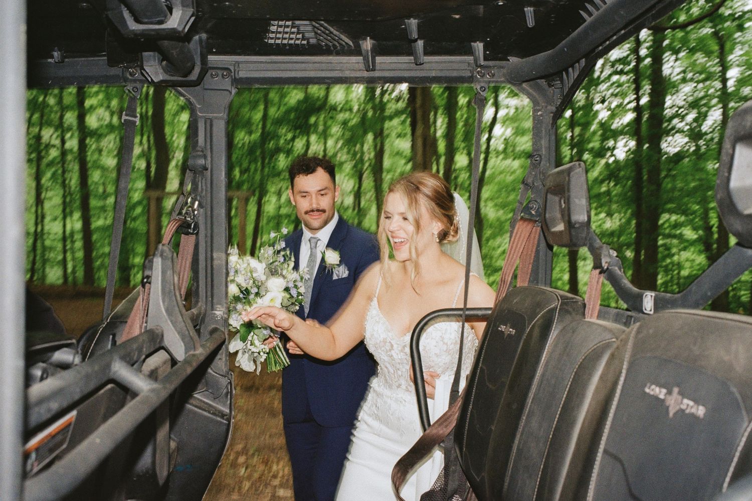 Bride and groom standing together inside a rustic vehicle surrounded by lush green forest trees on their wedding day.