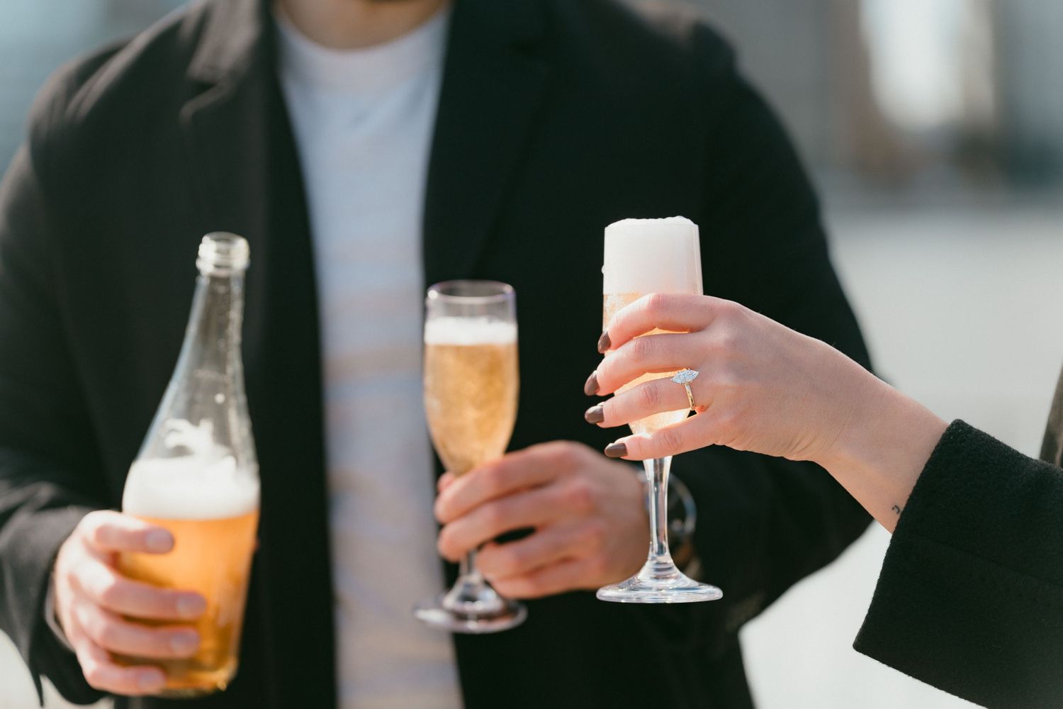 Close-up of an engaged couple holding champagne flutes and a bottle outdoors.
