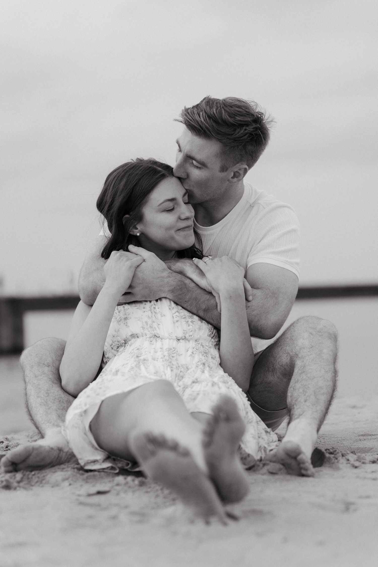 Black and white photo of a couple sitting on a beach, man kissing woman on the cheek as she smiles.