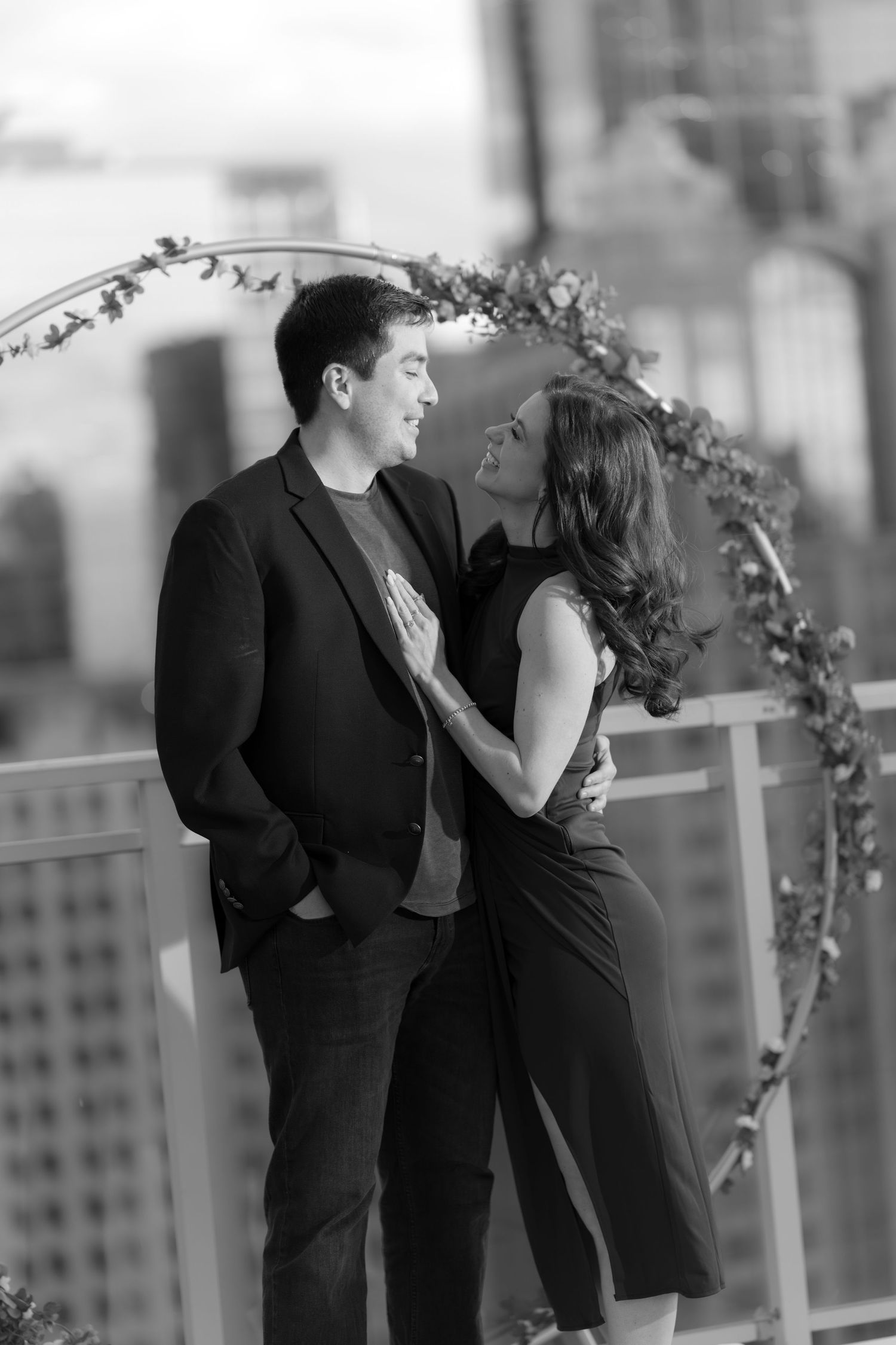 Black and white portrait of smiling couple standing together in front of circular floral arch on rooftop.