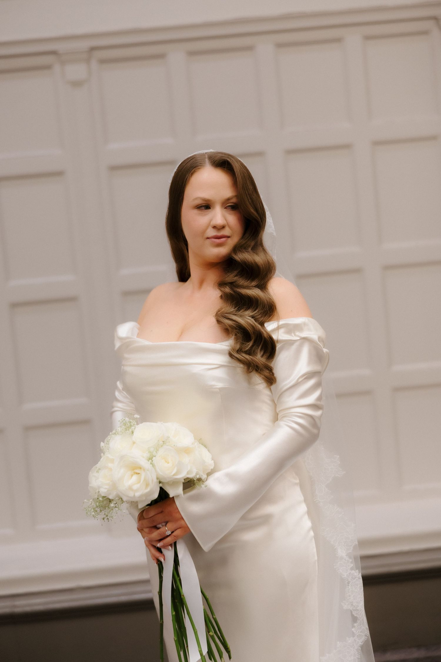 Elegant bride in white off-shoulder satin gown holding white rose bouquet, wearing veil, posing before white paneled wall.
