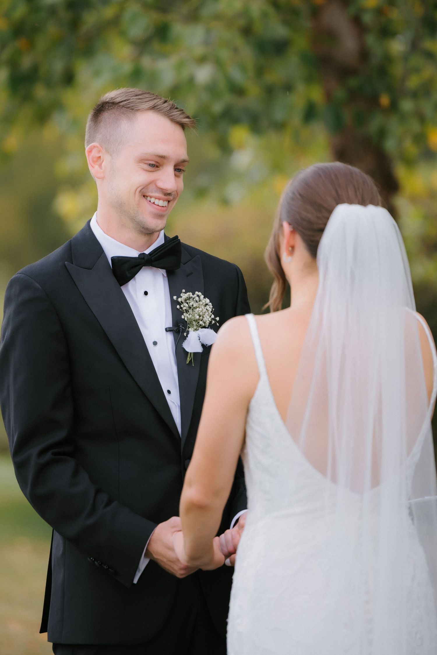 Joyful groom in black tuxedo holds brides hands and smiles during intimate outdoor wedding ceremony.
