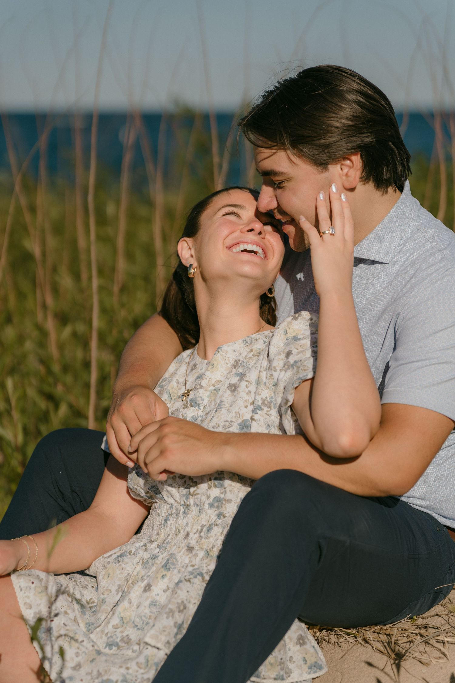 Couple sitting together on sandy beach grass, laughing and sharing a romantic moment during golden hour engagement session.