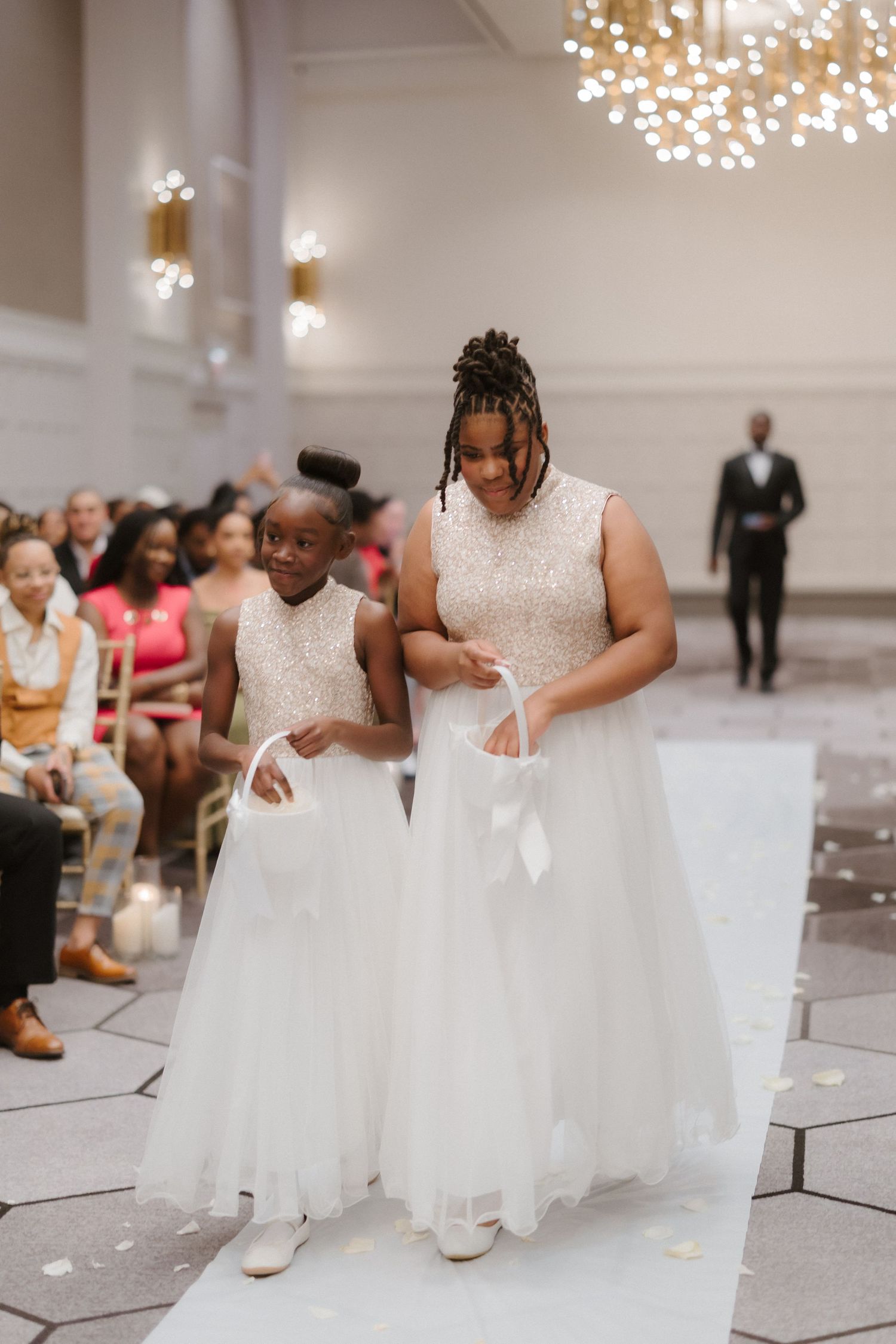 Two flower girls in white dresses walk down the wedding aisle carrying baskets during an elegant indoor ceremony.