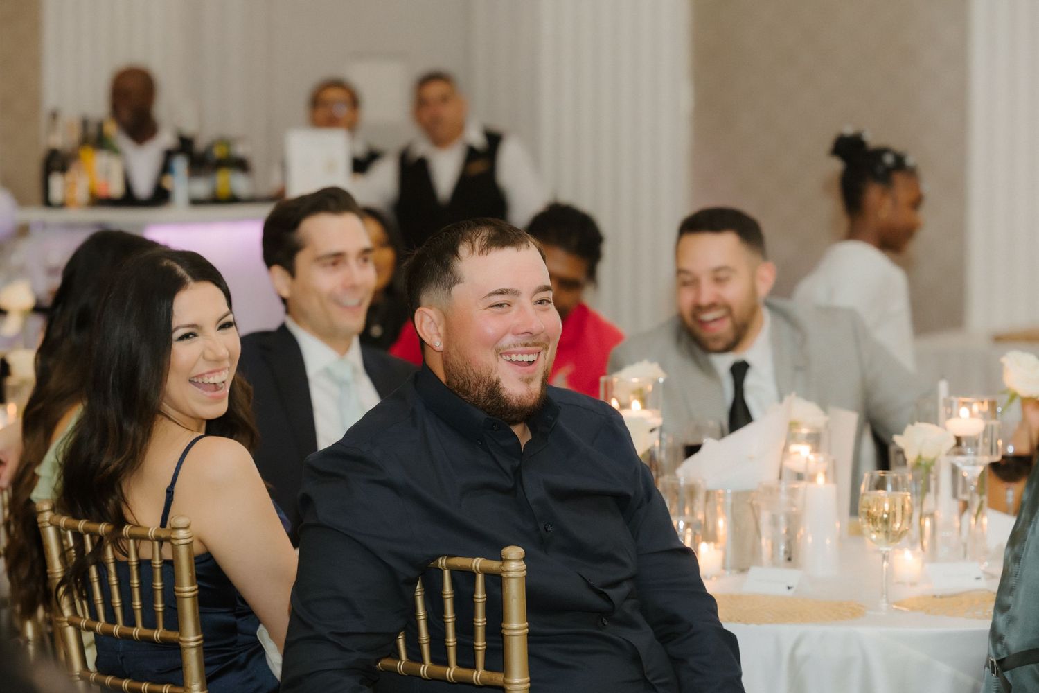 Guests laughing and enjoying themselves at an elegant wedding reception with candles and gold chairs.