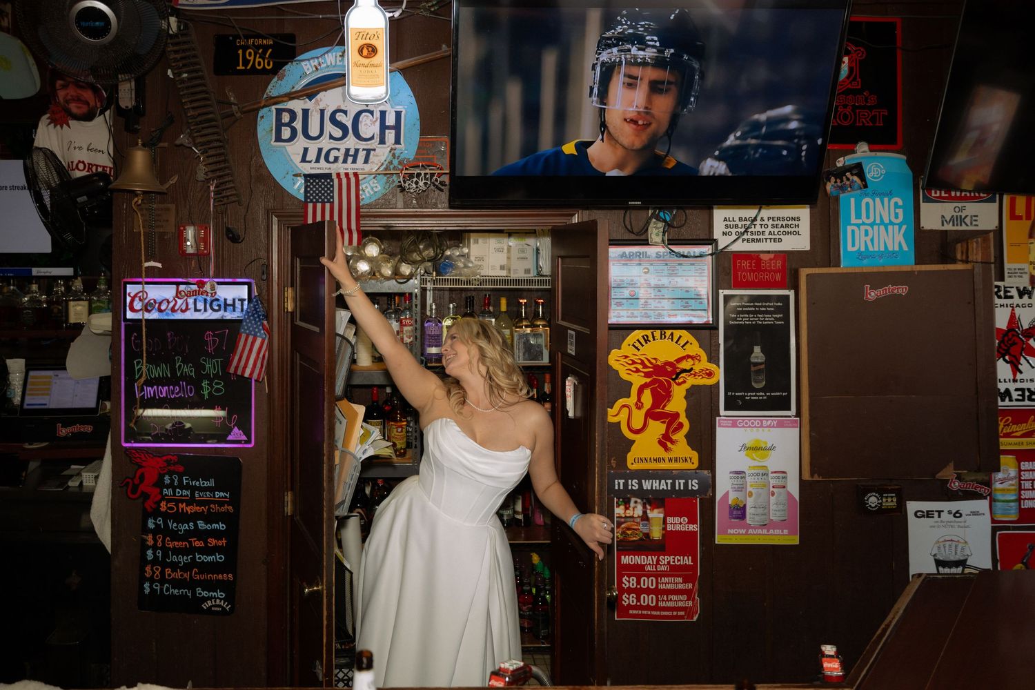 Bride in white dress reaching up near Busch Light sign in a bar decorated with posters and neon signs.