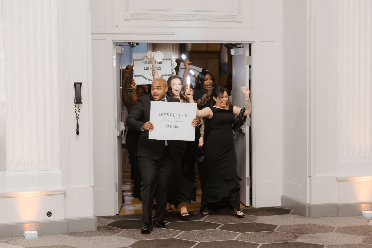 Wedding party exits venue holding a sign reading lets get the party started, dressed in elegant black attire.