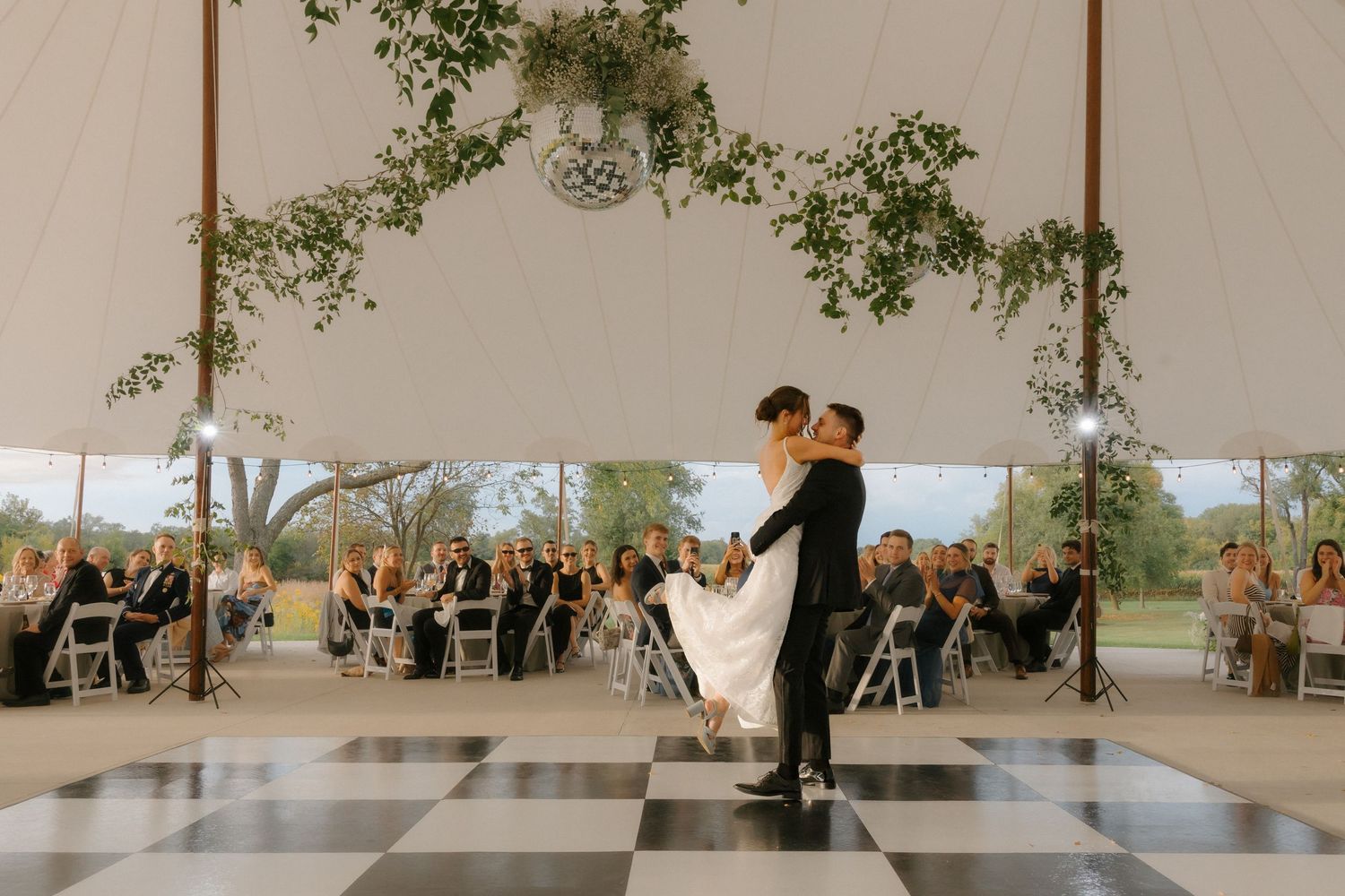 Groom lifts bride joyfully on a black and white checkered dance floor as guests watch their first dance.