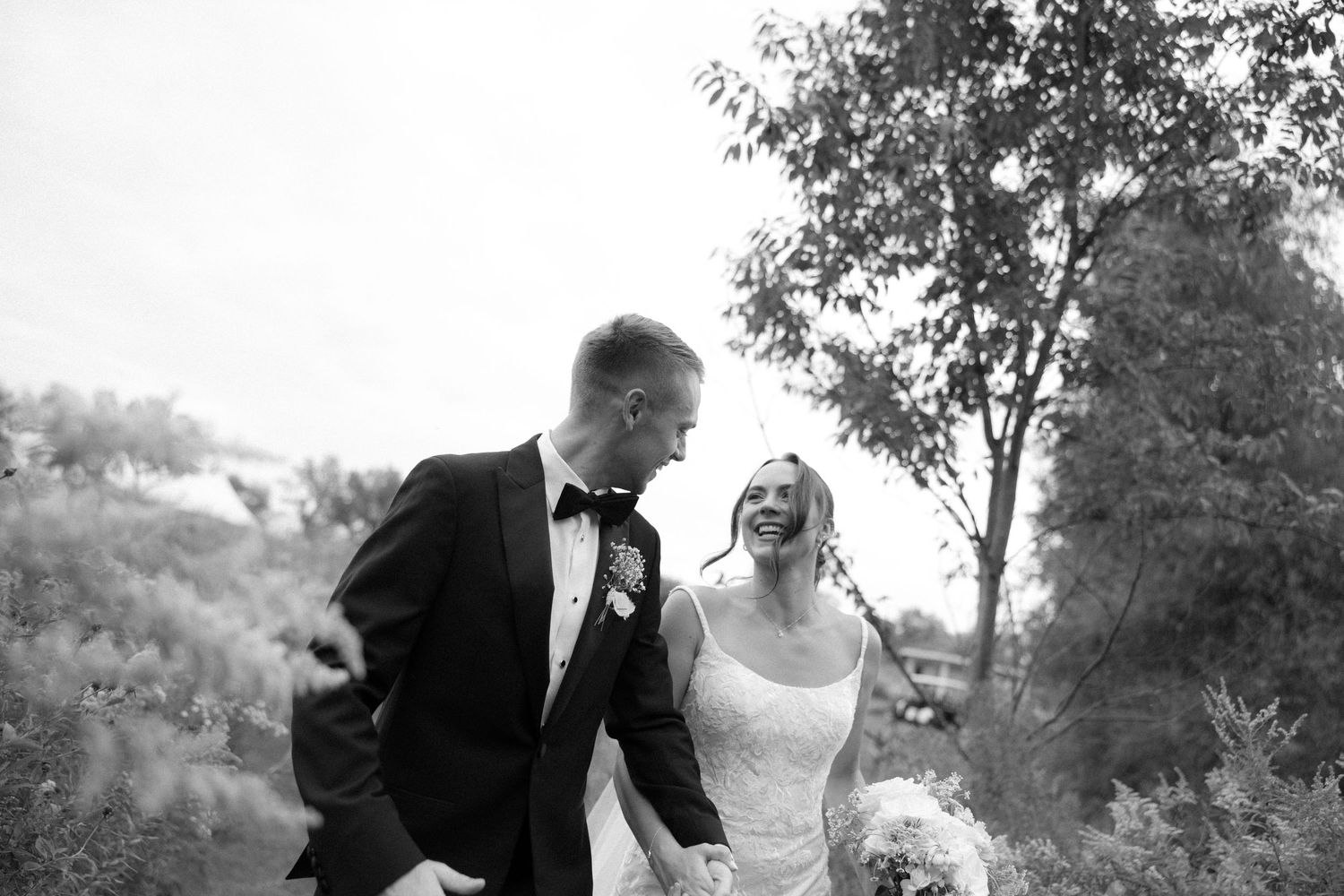 Black and white photo of bride and groom laughing while walking hand in hand through lush greenery outdoors.