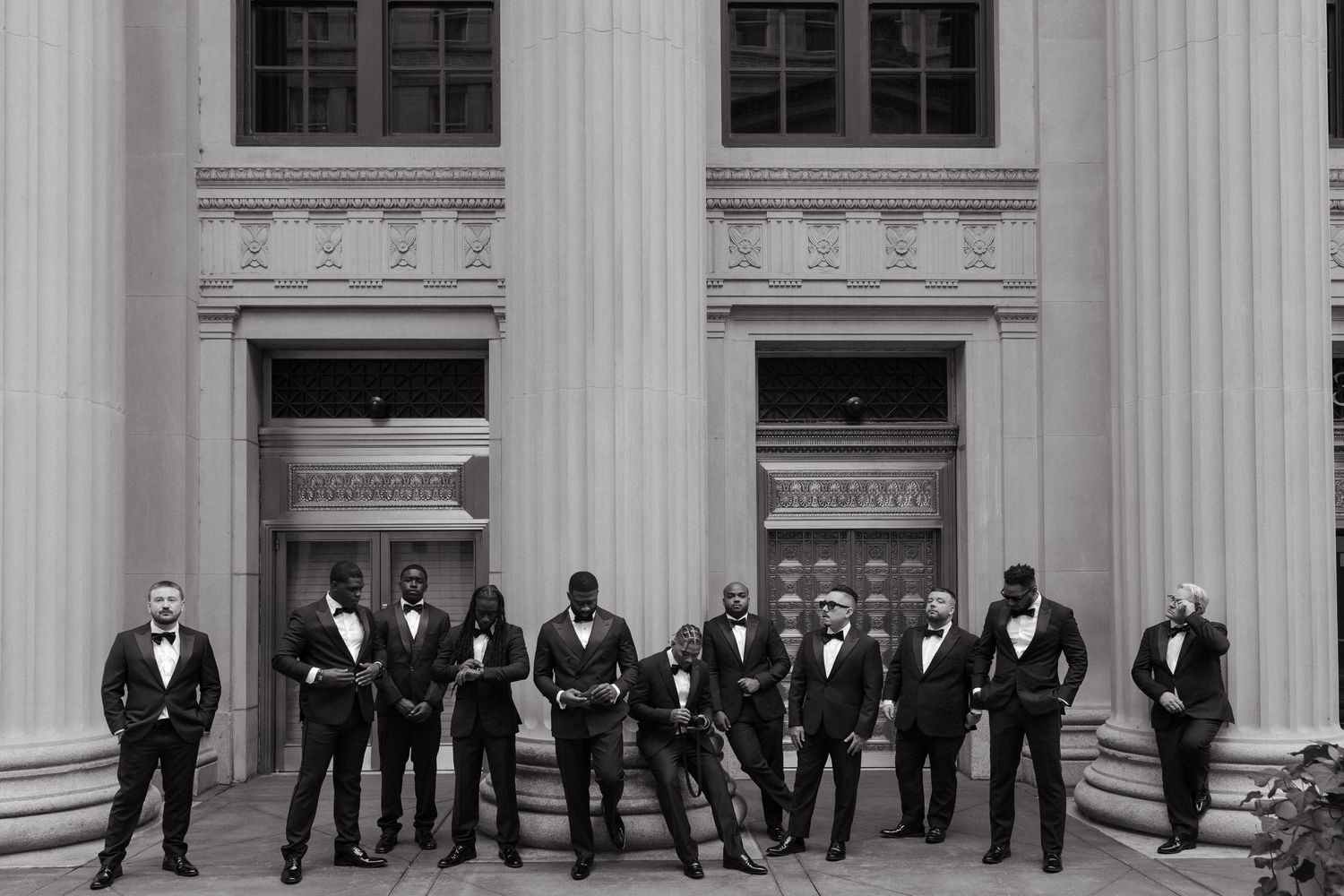 Groomsmen in black tuxedos posing on steps of a neoclassical building with large columns in black and white photo.
