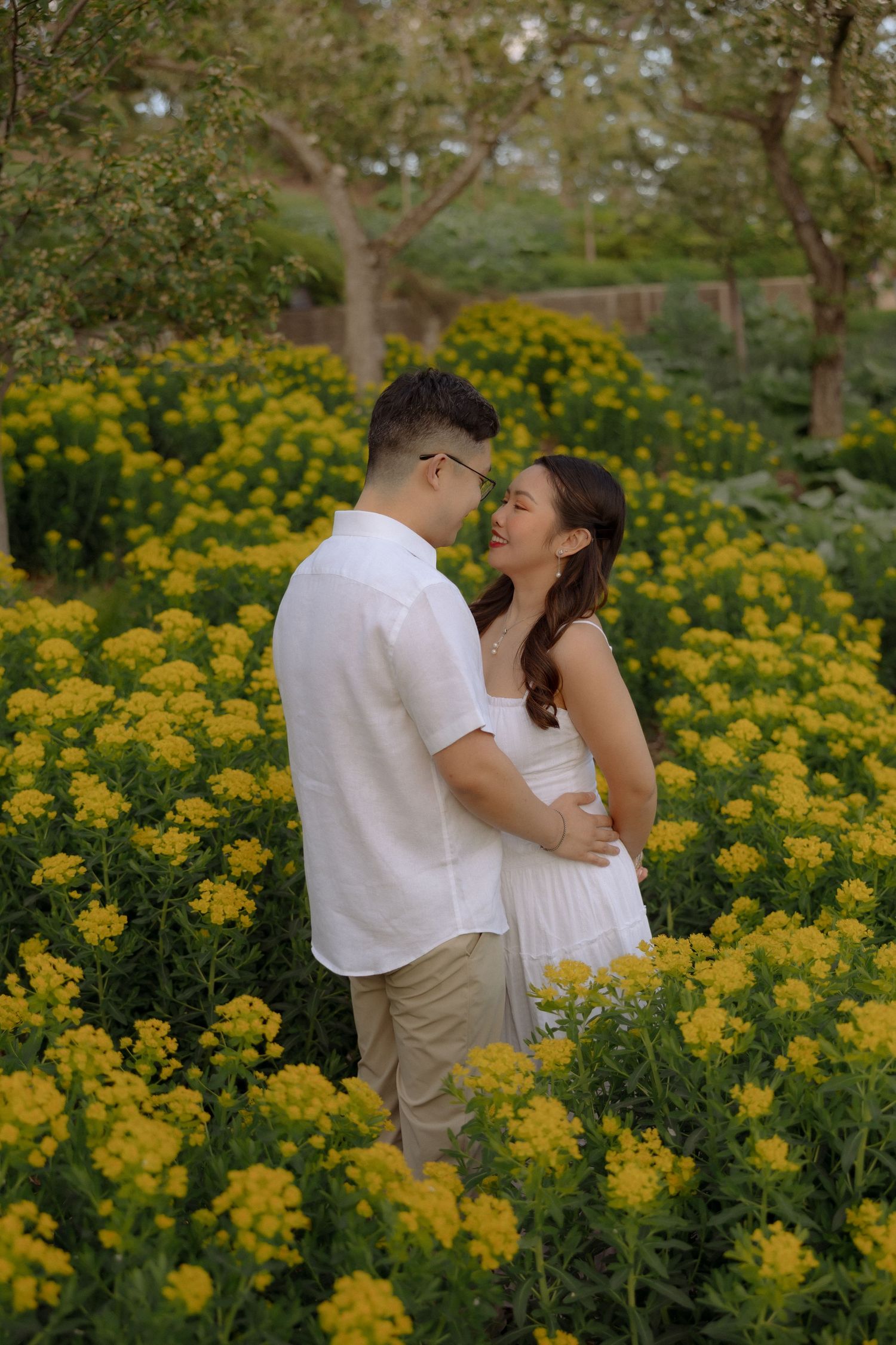 Couple embracing romantically among vibrant yellow flowers in a garden, wearing white outfits, sharing an intimate moment.