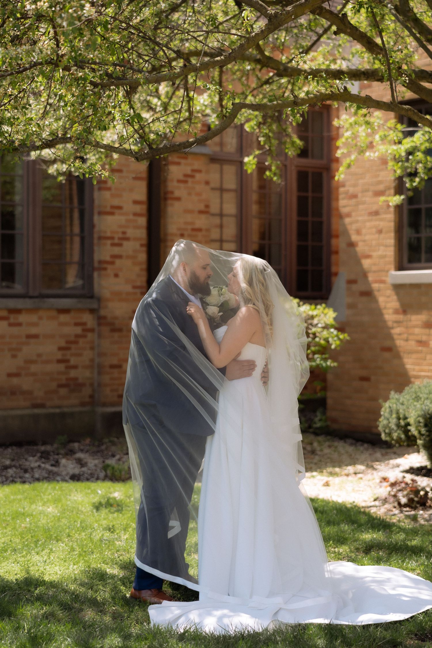 Bride and groom sharing a kiss under a flowing veil outdoors near a brick building on their wedding day.