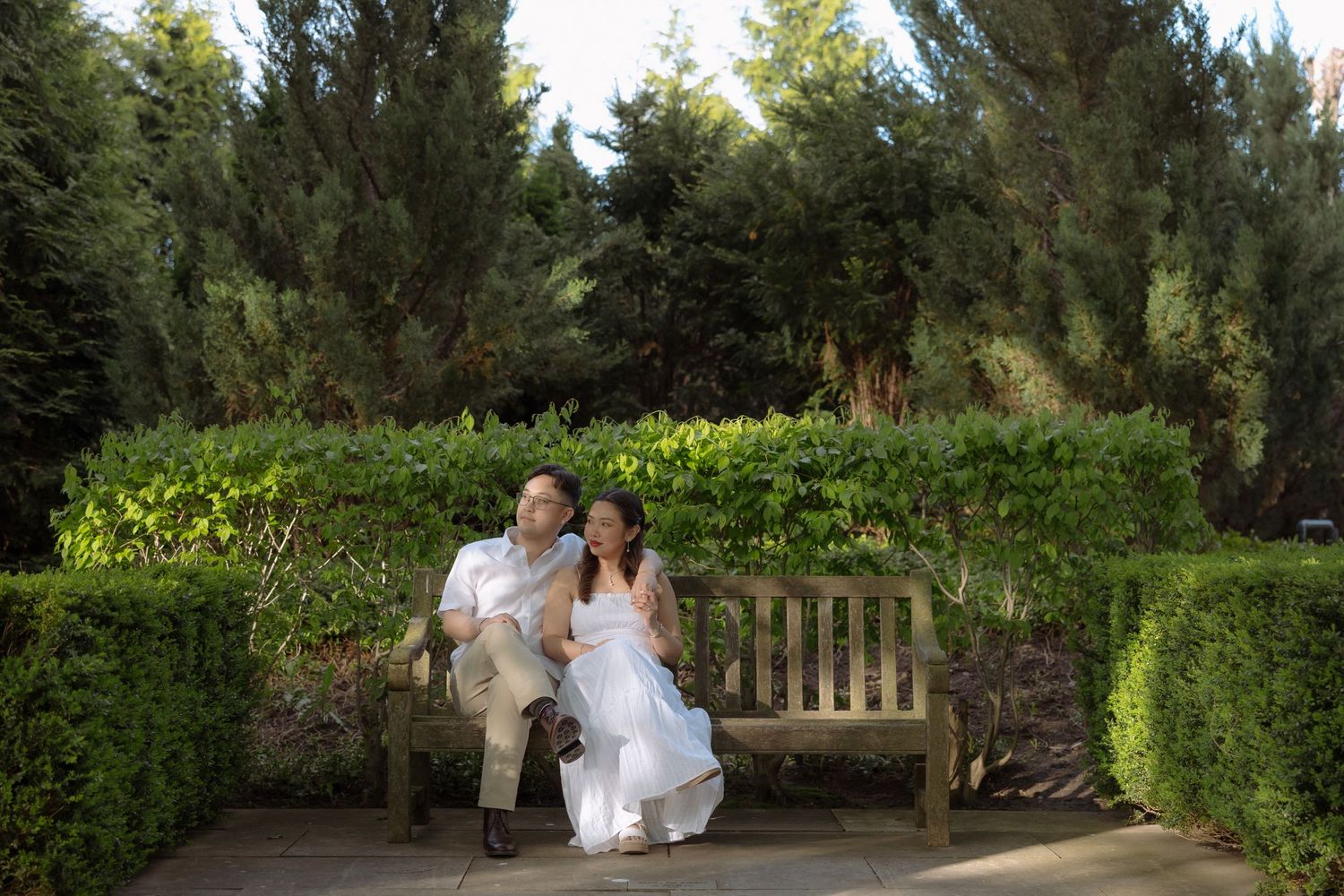 A couple in wedding attire sits on a wooden bench surrounded by lush green hedges and tall trees in a garden.
