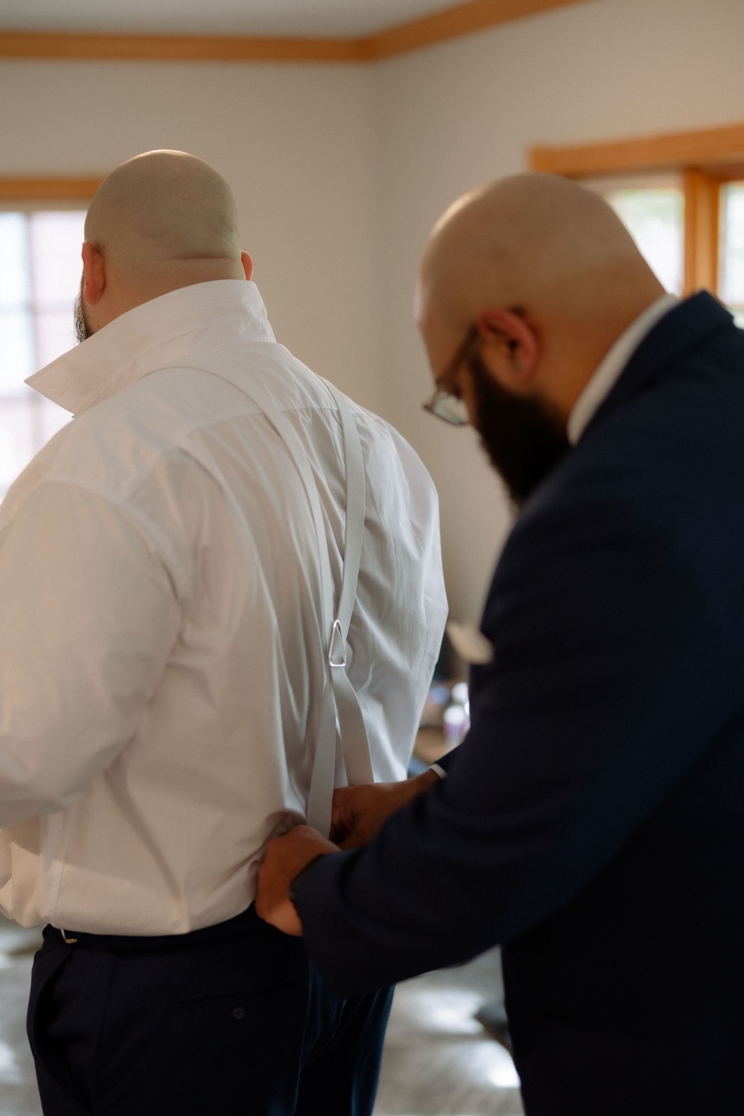 Two bald men in formal attire, one in a navy suit helping the other button a white dress shirt indoors.