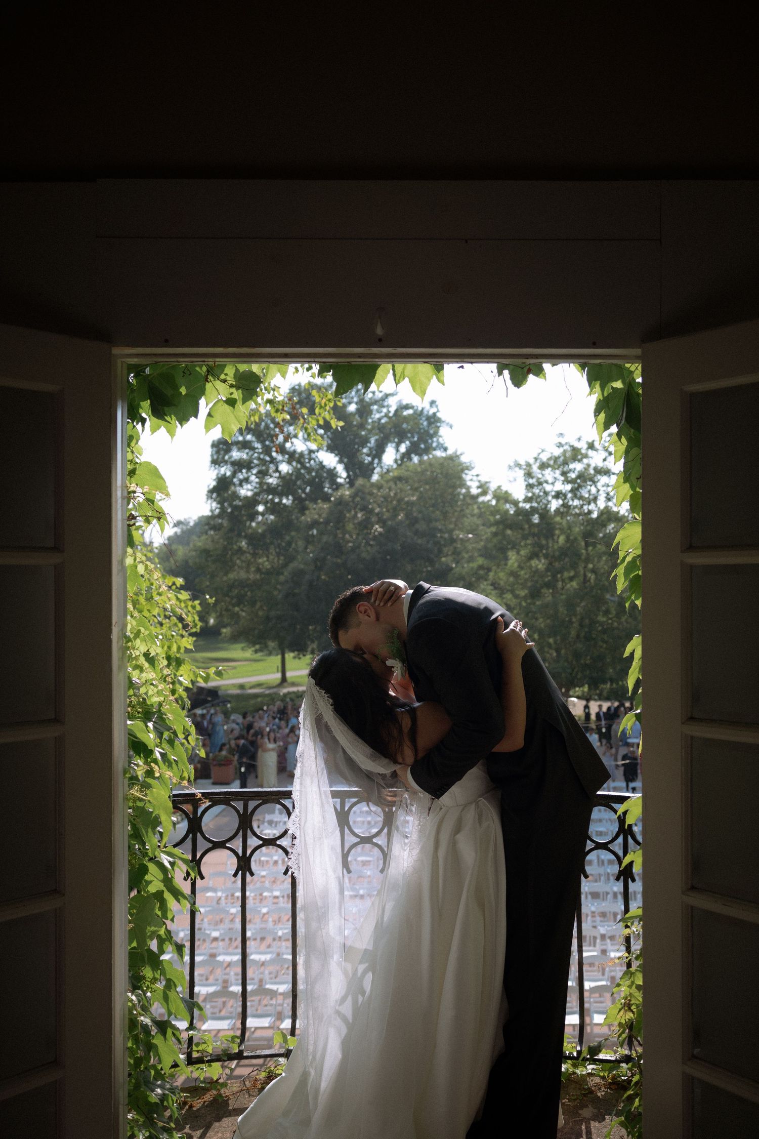 Bride and groom share a romantic first kiss framed by an open doorway with lush green vines at their wedding.