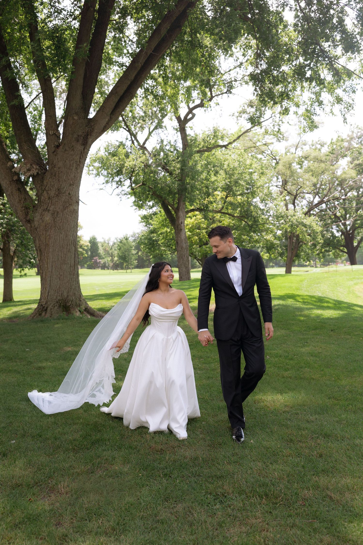Bride in white gown and groom in black tuxedo walk hand in hand across lush green park lawn.
