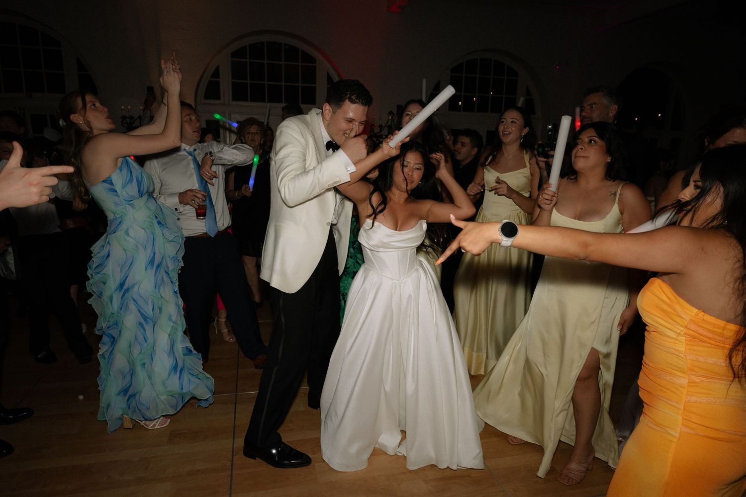 Bride and groom celebrate on crowded wedding reception dance floor with glowing light sticks.