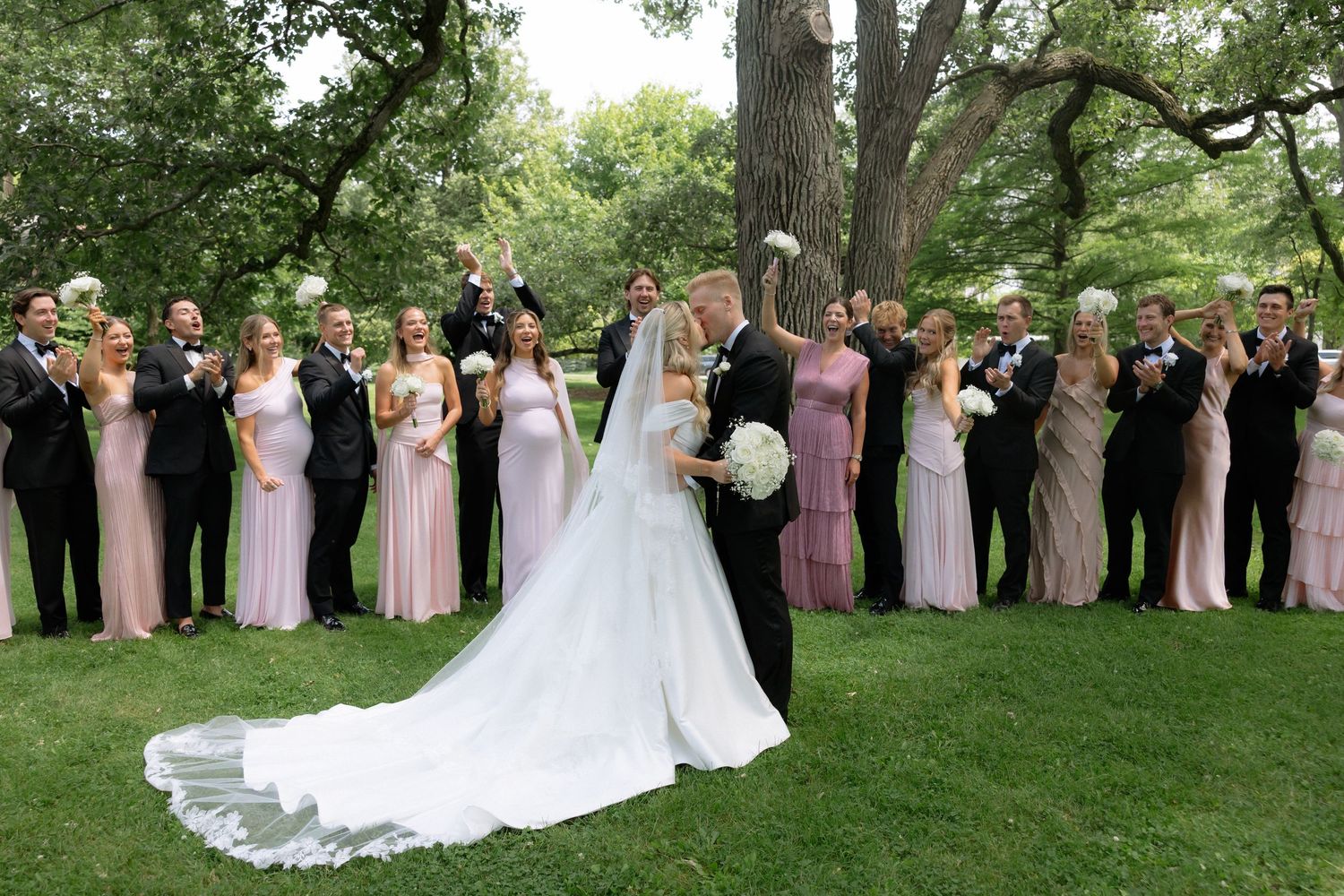 Wedding party celebrating outdoors under trees with bridesmaids in pink dresses and groomsmen in tuxedos.