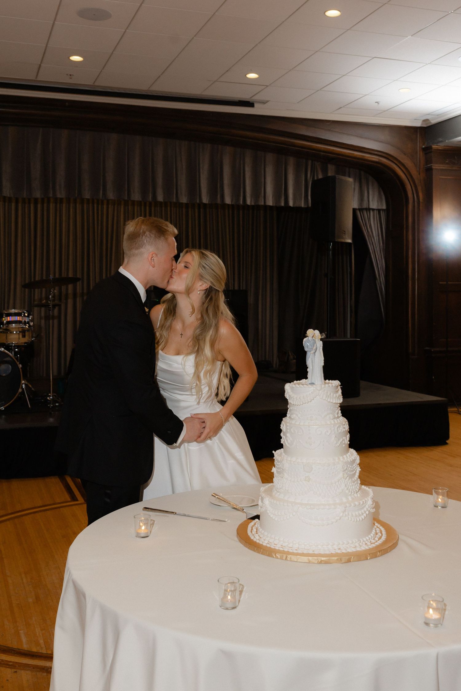 Bride and groom kissing beside elegant five-tier white wedding cake at indoor reception.