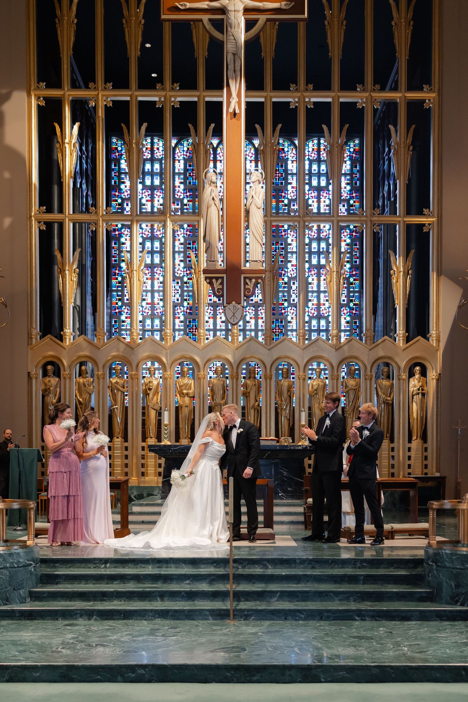Bride and groom exchange vows at ornate church altar with stained glass windows and golden decor.