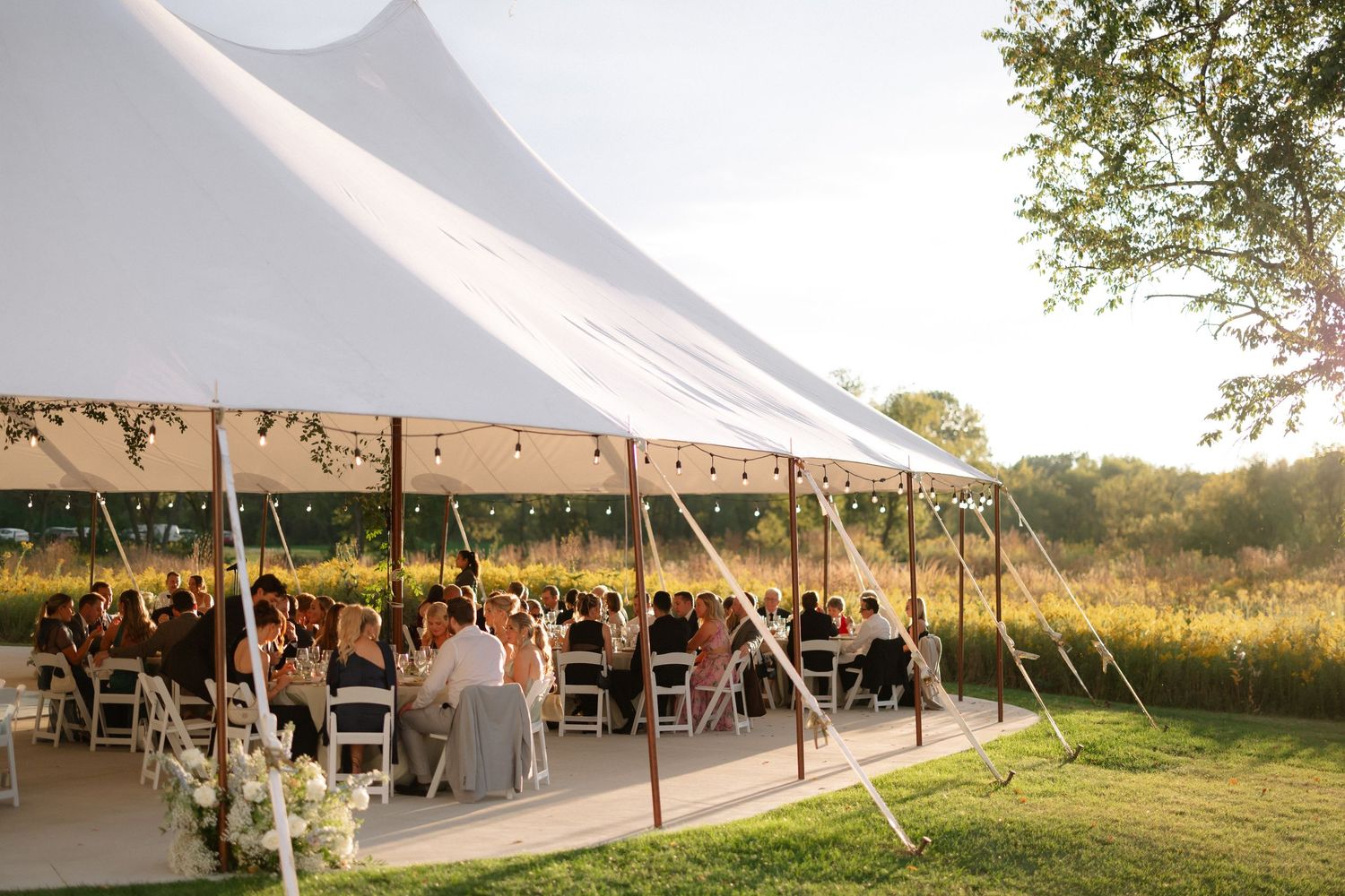 Elegant outdoor wedding reception under a white sailcloth tent surrounded by lush greenery at golden hour.