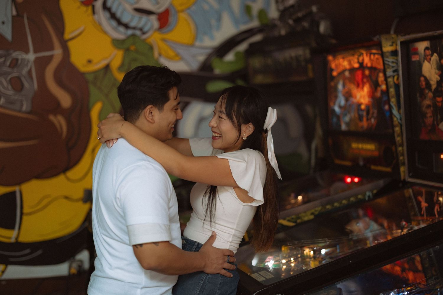 Couple embracing and smiling at each other in a retro arcade bar with colorful pinball machines and mural artwork.