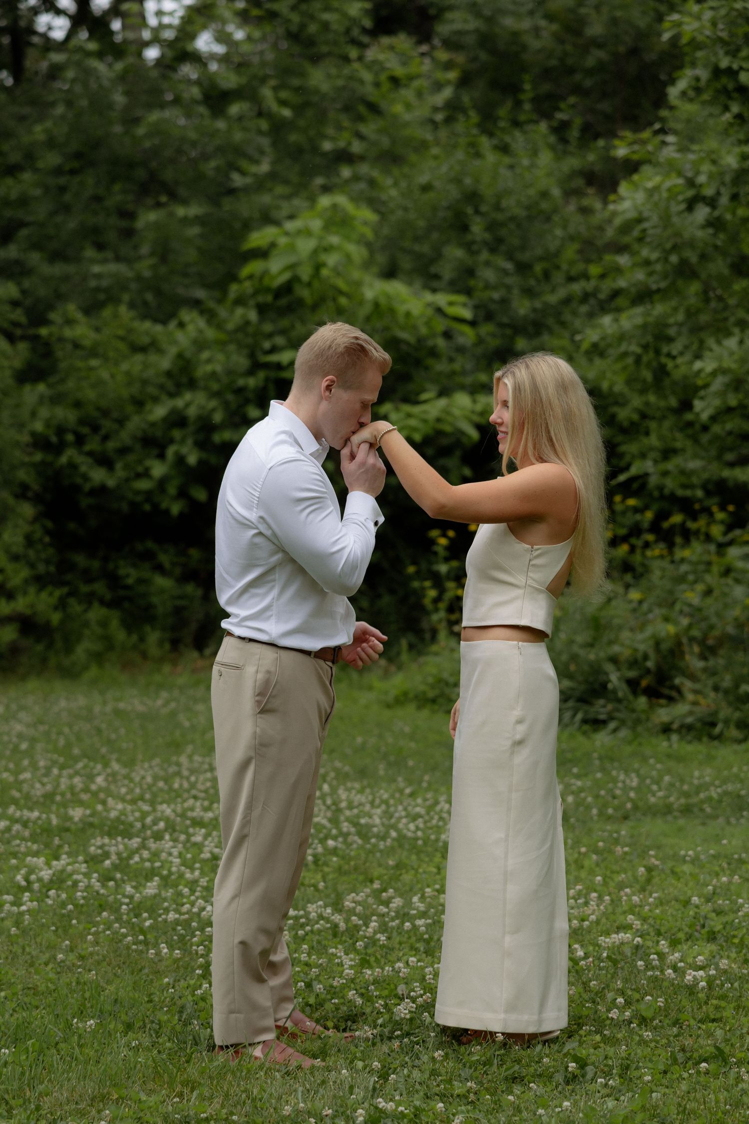 Man kissing woman's hand while couple stands in flower-dotted meadow during romantic engagement photo shoot.