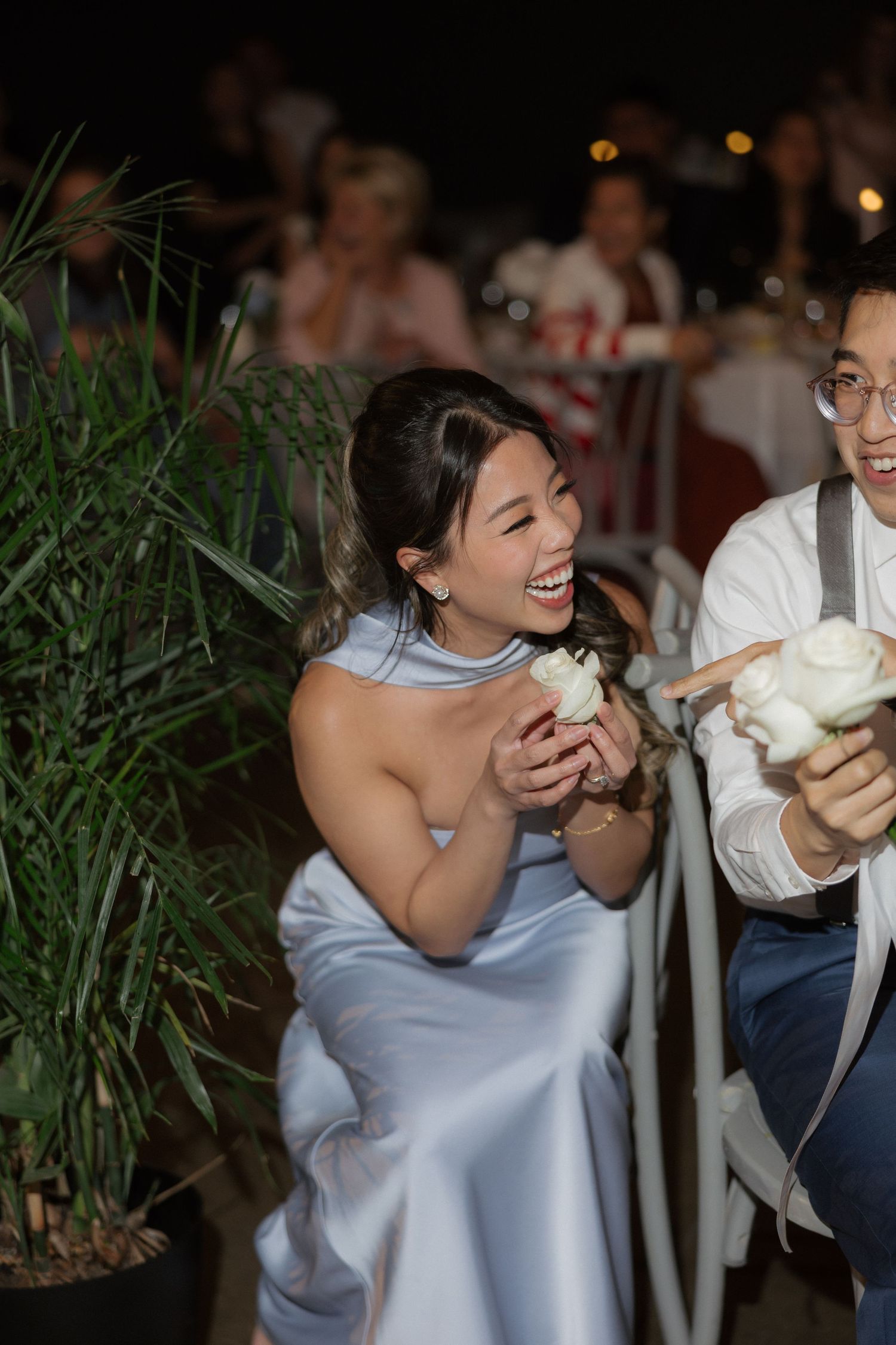 Laughing bridesmaid in light blue dress holding white flower at outdoor wedding reception.