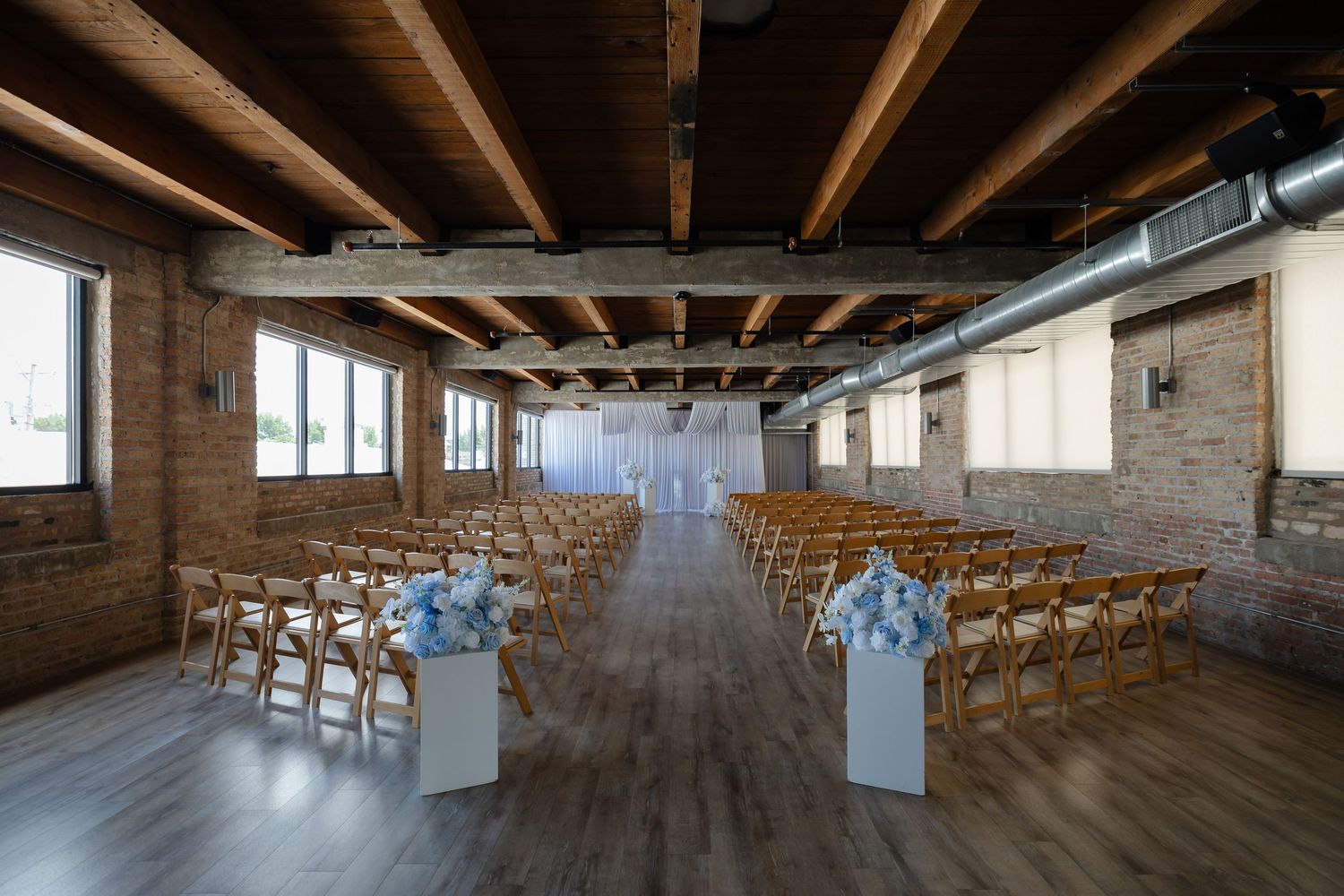 Elegant wedding ceremony setup in rustic brick venue with wooden chairs, floral arrangements, and exposed beam ceiling.