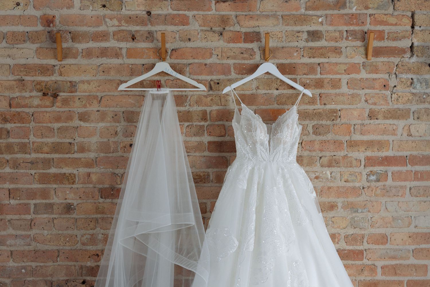 White lace wedding dress and veil hanging on wooden hangers against a rustic brick wall.