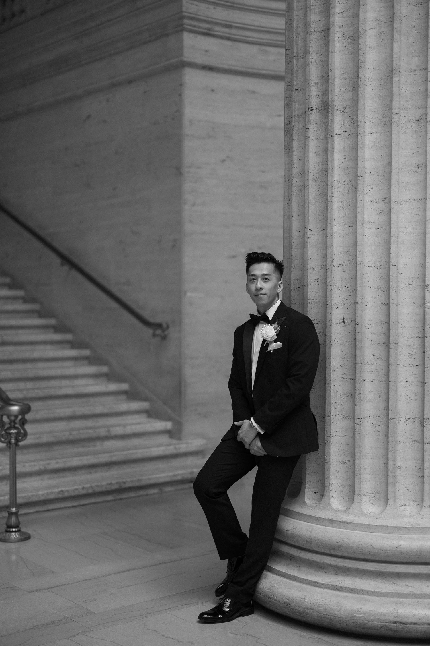 Dapper groom in black tuxedo leans against classical column in black and white wedding portrait.
