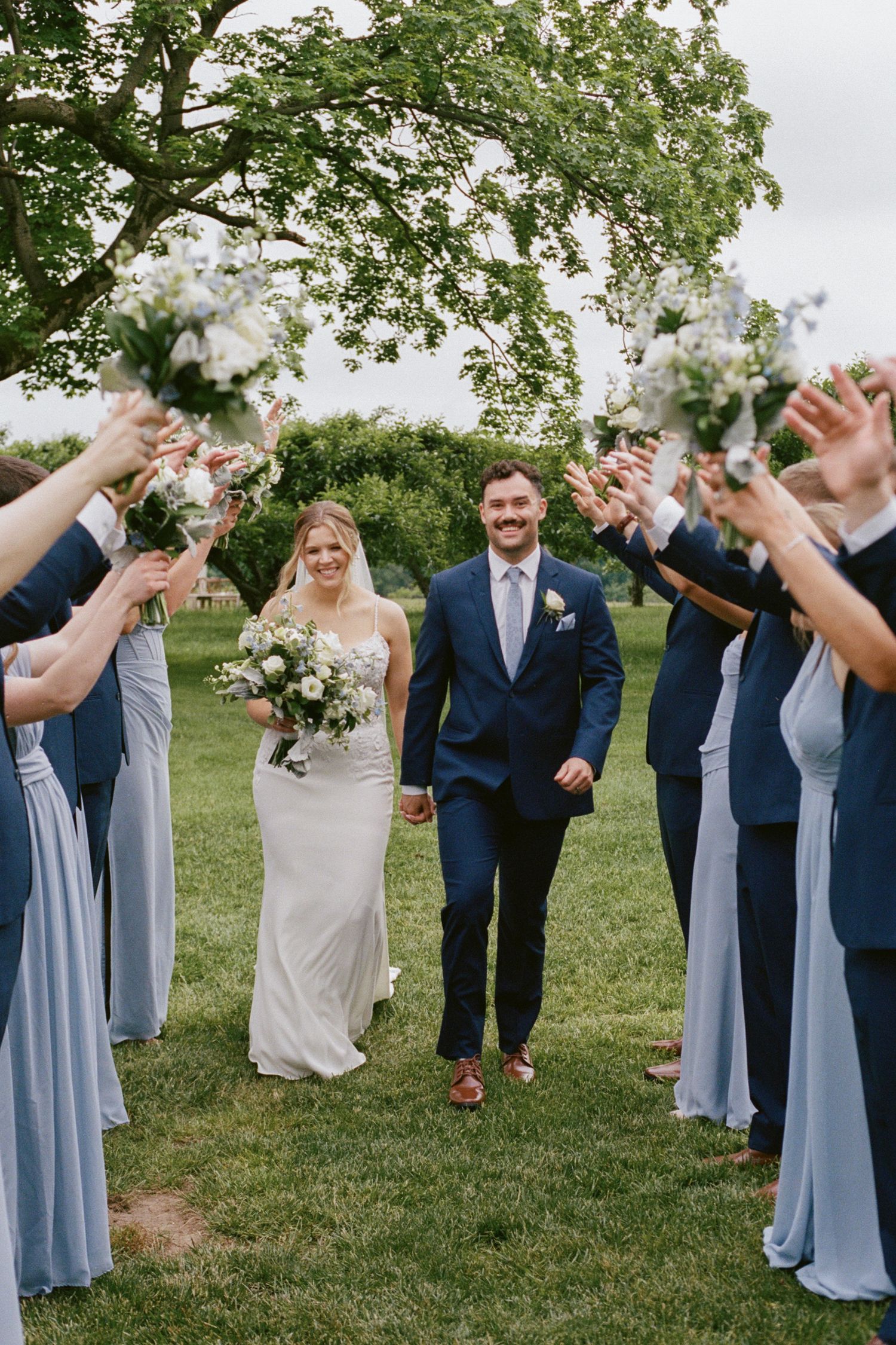 Bride and groom walk through an arch of raised bouquets, flanked by bridesmaids in blue and groomsmen in navy suits.
