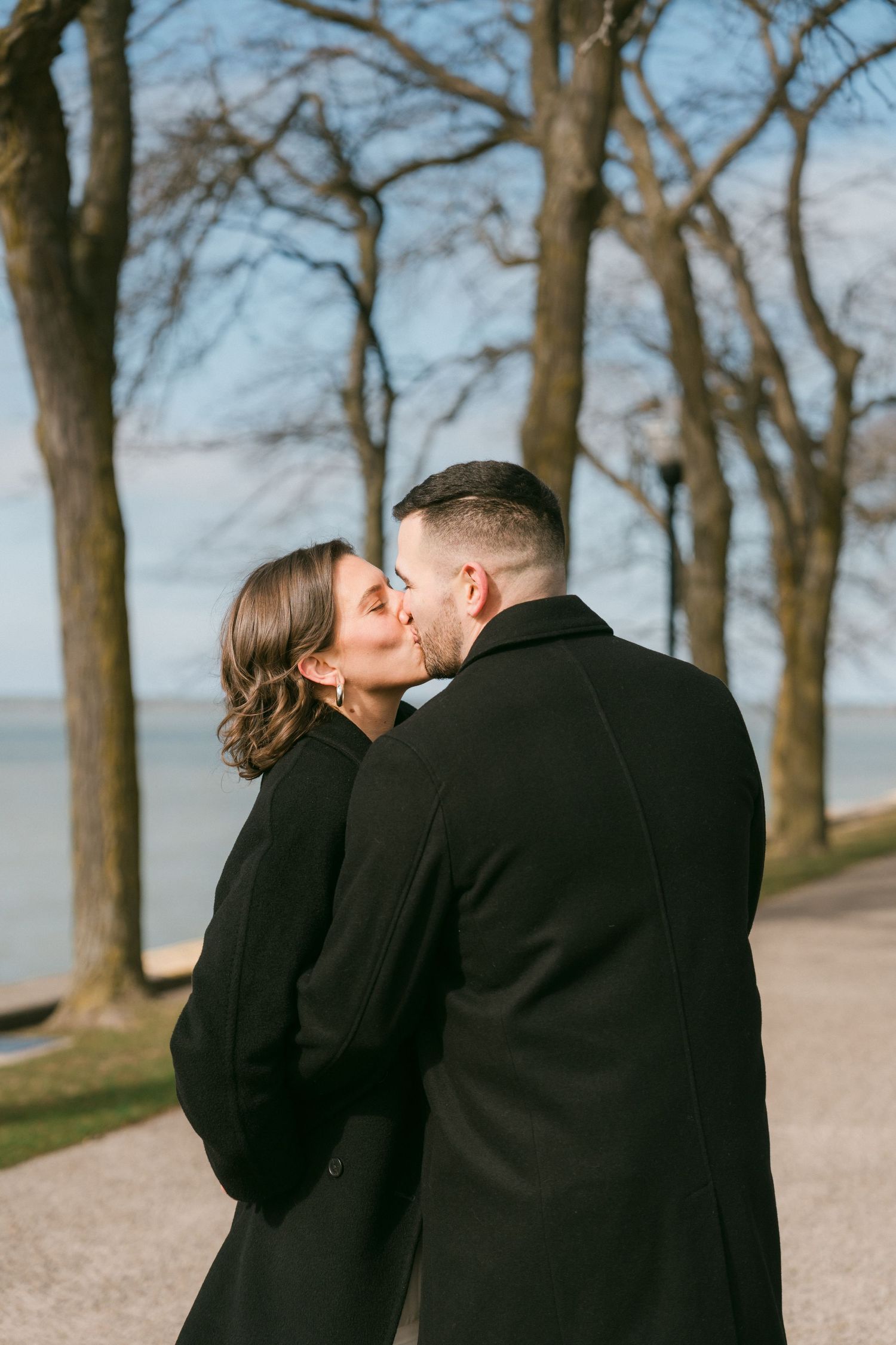 Romantic couple sharing a tender kiss among bare winter trees near Chicago lakefront waterfront path.