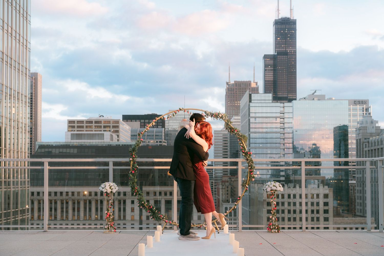 Newly engaged couple embraces under circular floral arch on Chicago rooftop with Willis Tower in background.