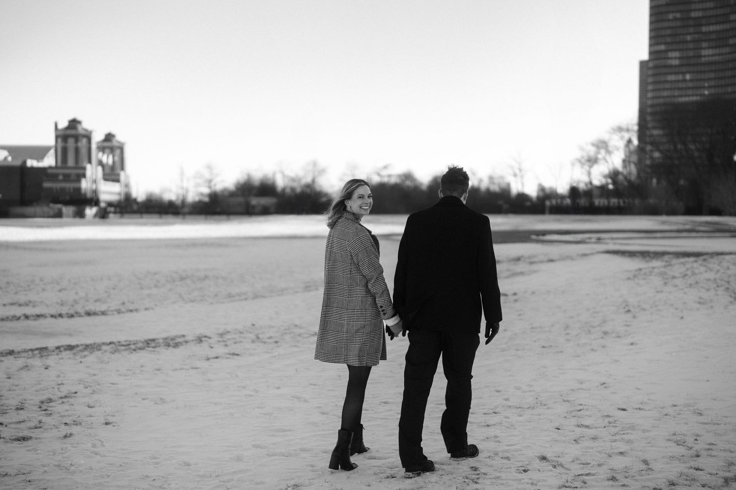A couple holds hands while walking away on a snowy landscape at dusk in black and white.