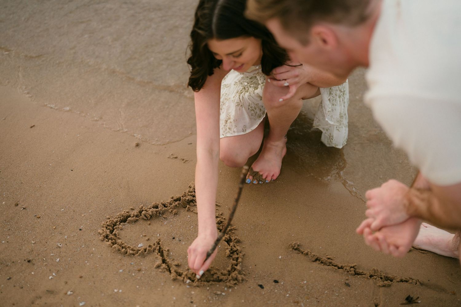 Couple crouching on wet sand drawing a heart shape together on a beach during their engagement session.