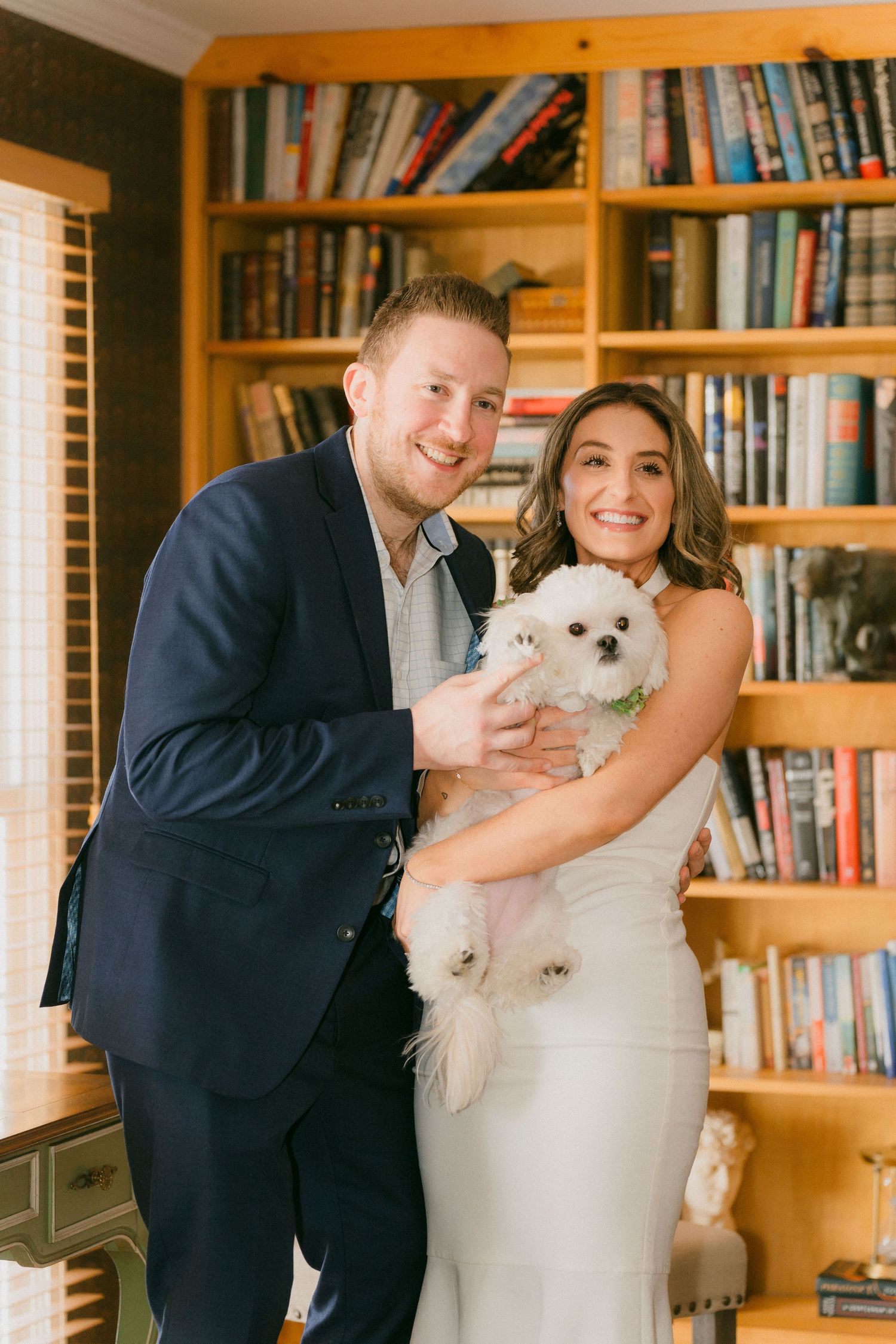 Happy newlywed couple holding their small white dog in front of a large bookshelf at home.