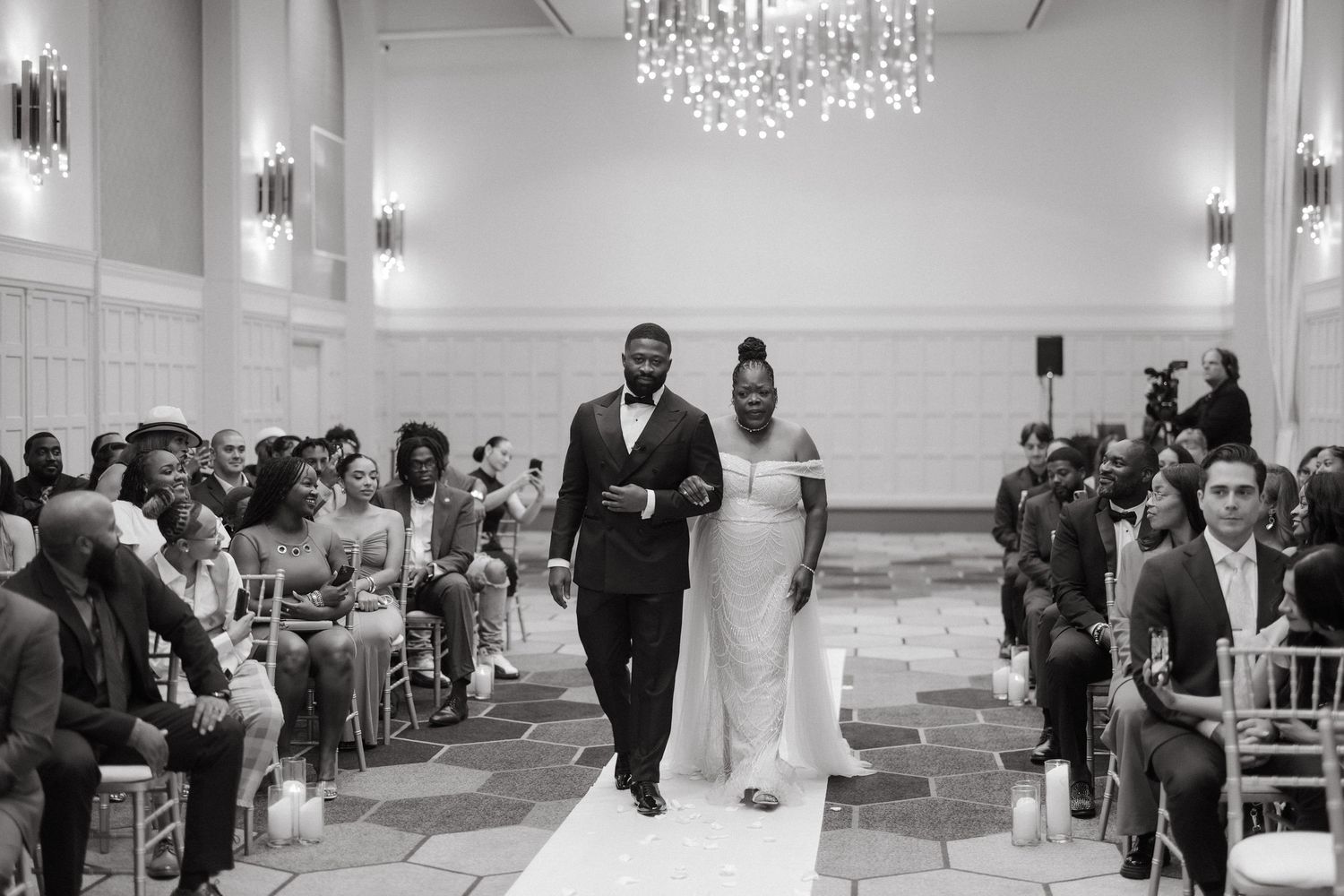 Black and white photo of a couple walking down the wedding aisle in an elegant ballroom with chandelier.