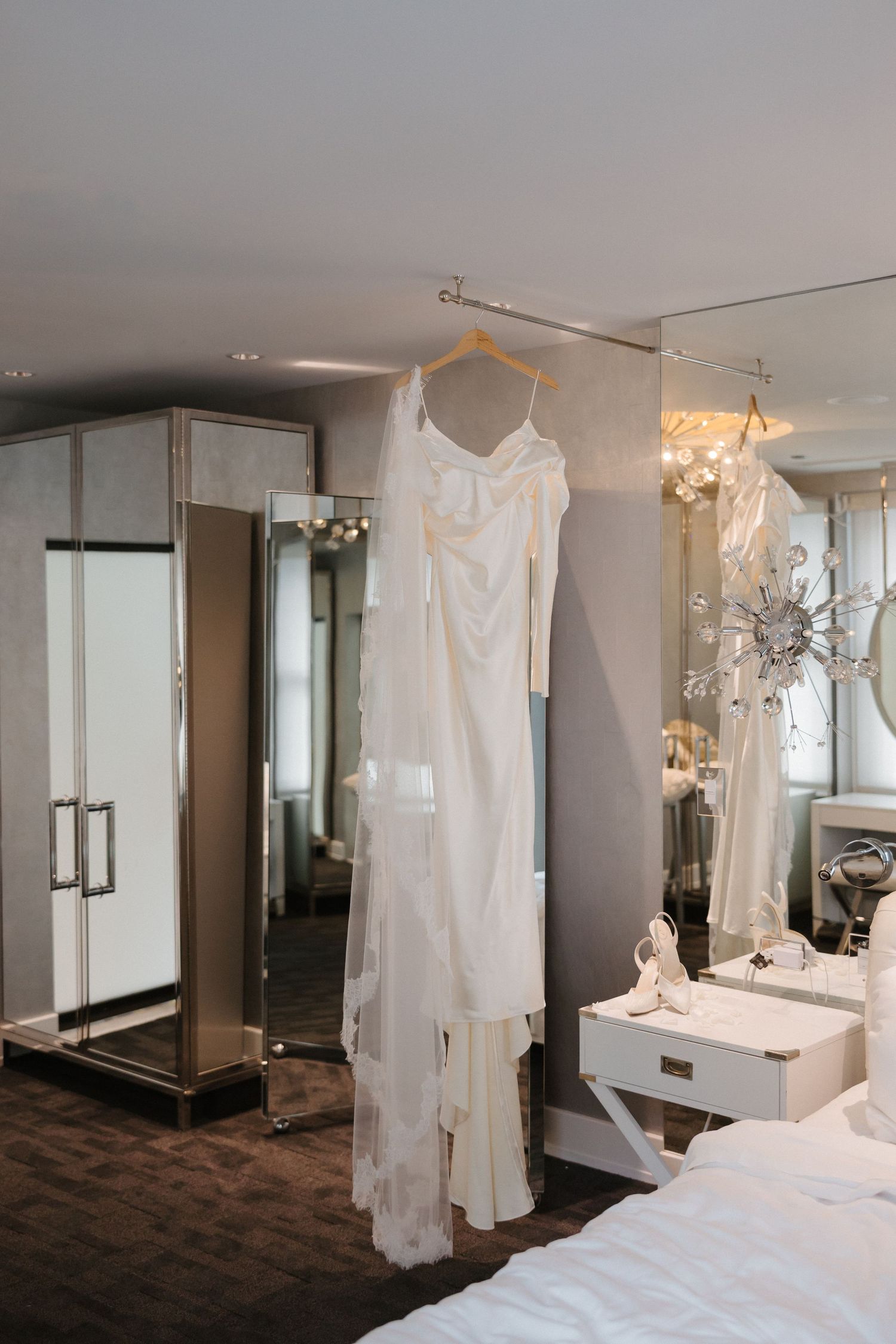 Elegant white bridal gown hanging on a hanger in a luxurious mirrored bedroom before the wedding ceremony.