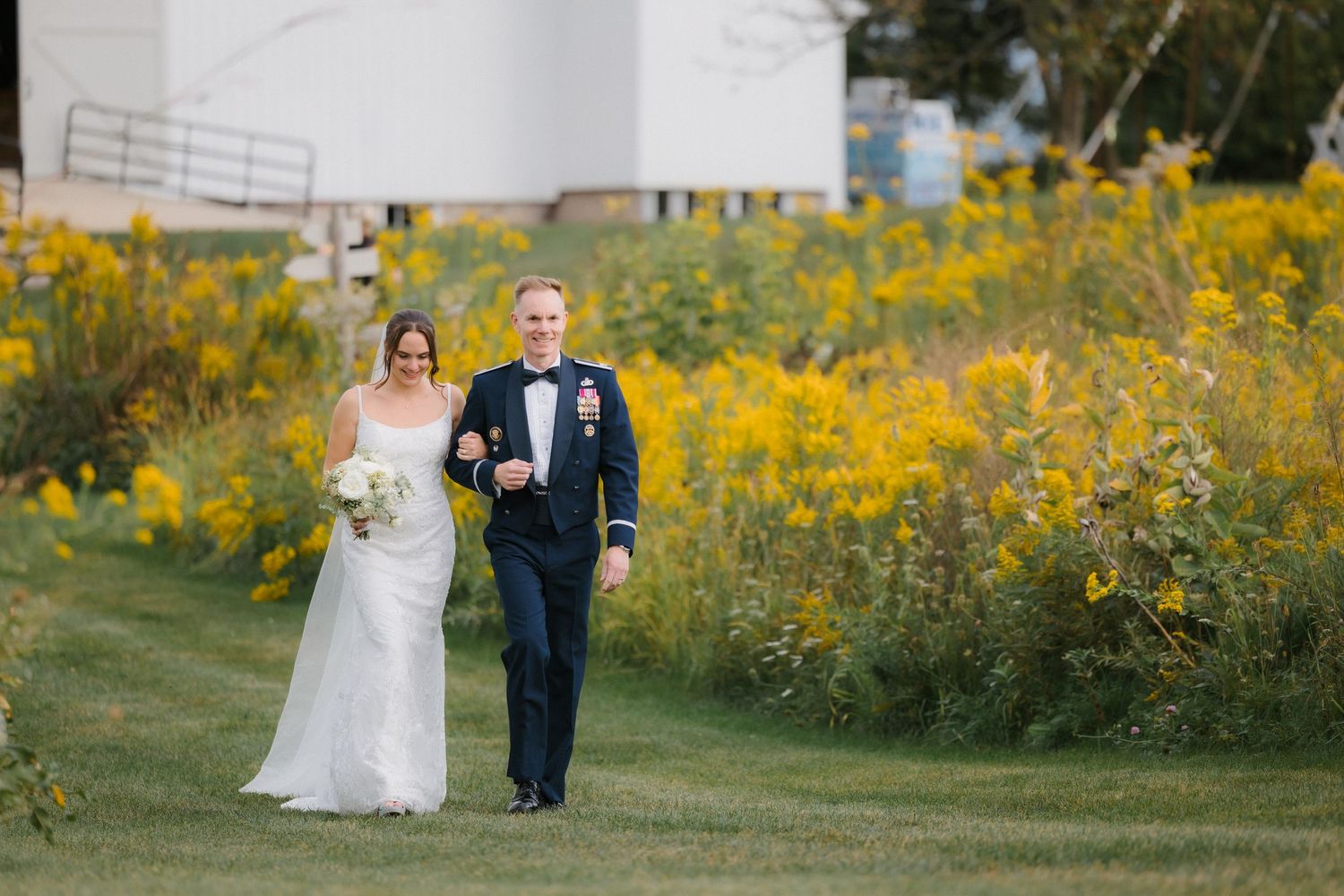 Bride in white gown and groom in military dress uniform walk arm-in-arm past vibrant yellow wildflowers.