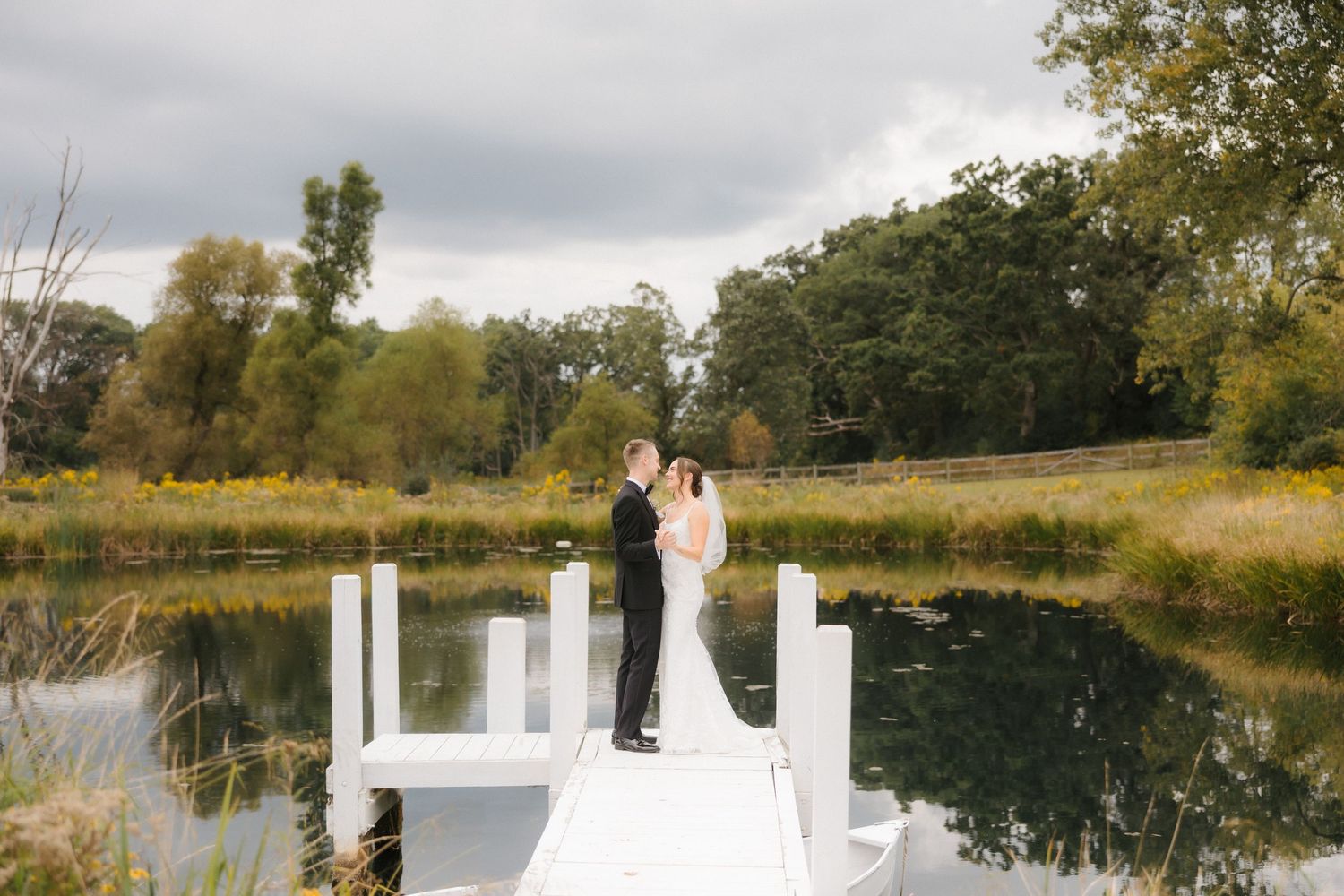 Bride and groom posing on a white dock with a tranquil lake and colorful autumn landscape reflected below.