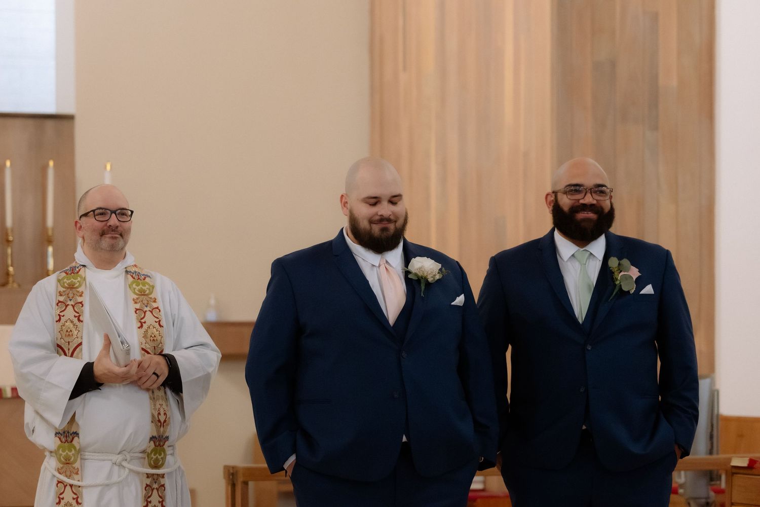 Two groomsmen in navy suits stand with a priest at a church wedding ceremony altar.